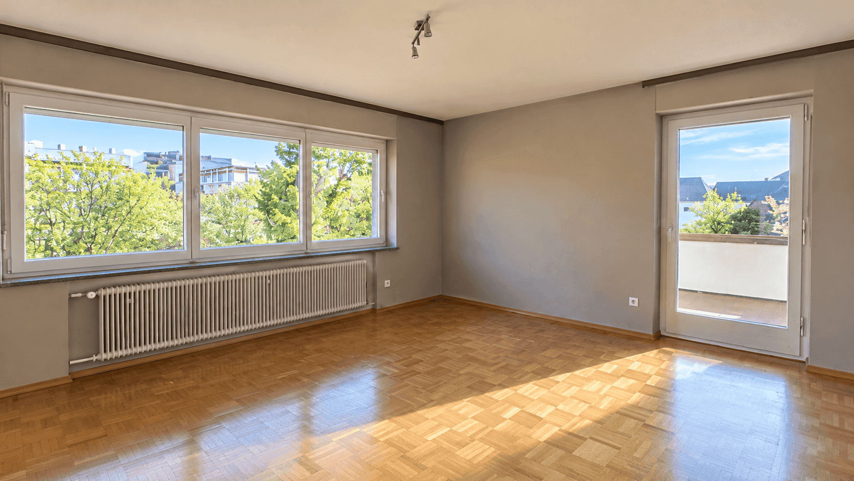 Leerer Raum mit Parkettboden, grauen Wänden, großen Fenstern und Zugang zu einem kleinen Balkon.