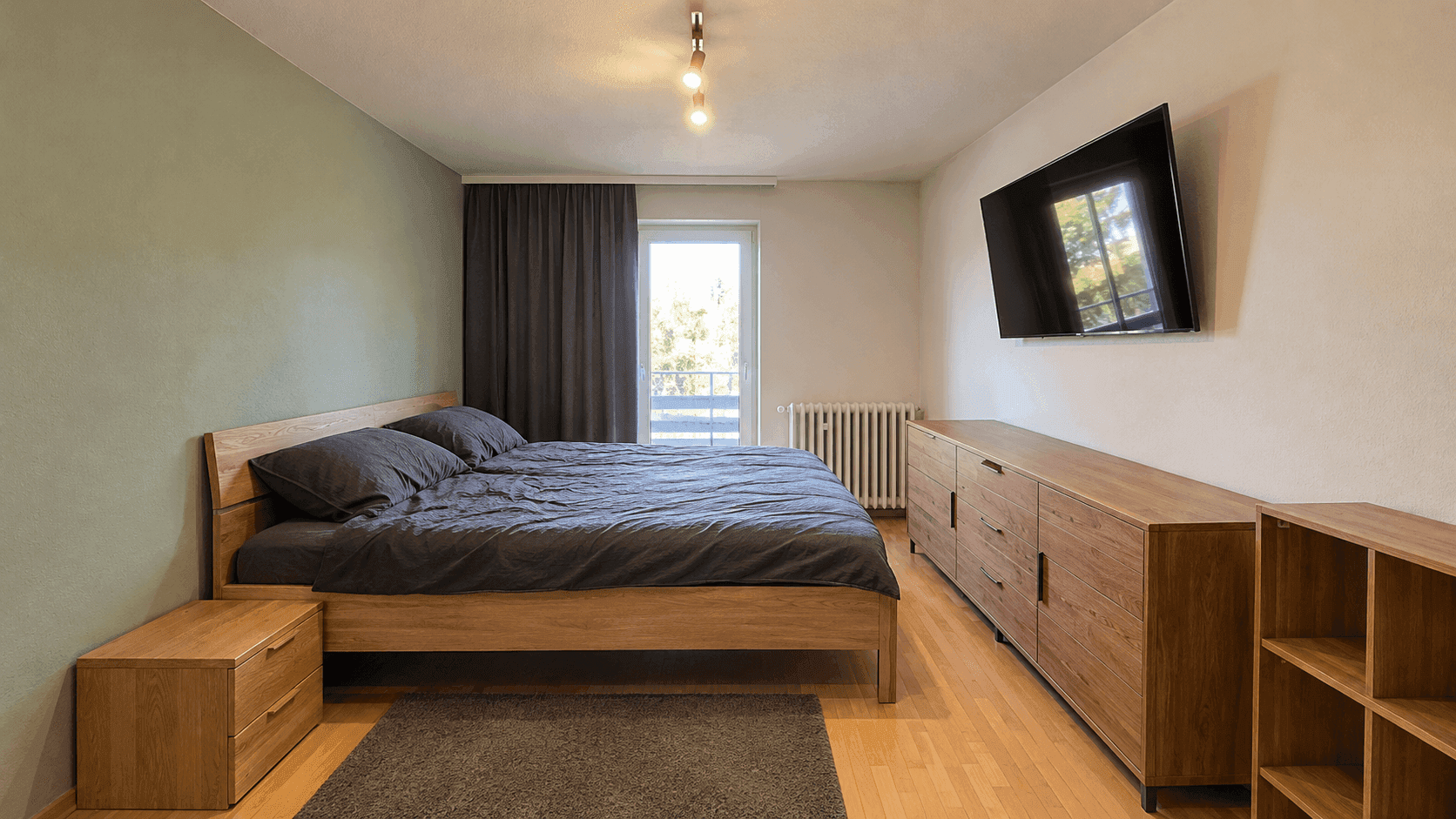 Schlafzimmer mit Holzbett, Parkettboden, Kommode und Zugang zu einem Balkon.