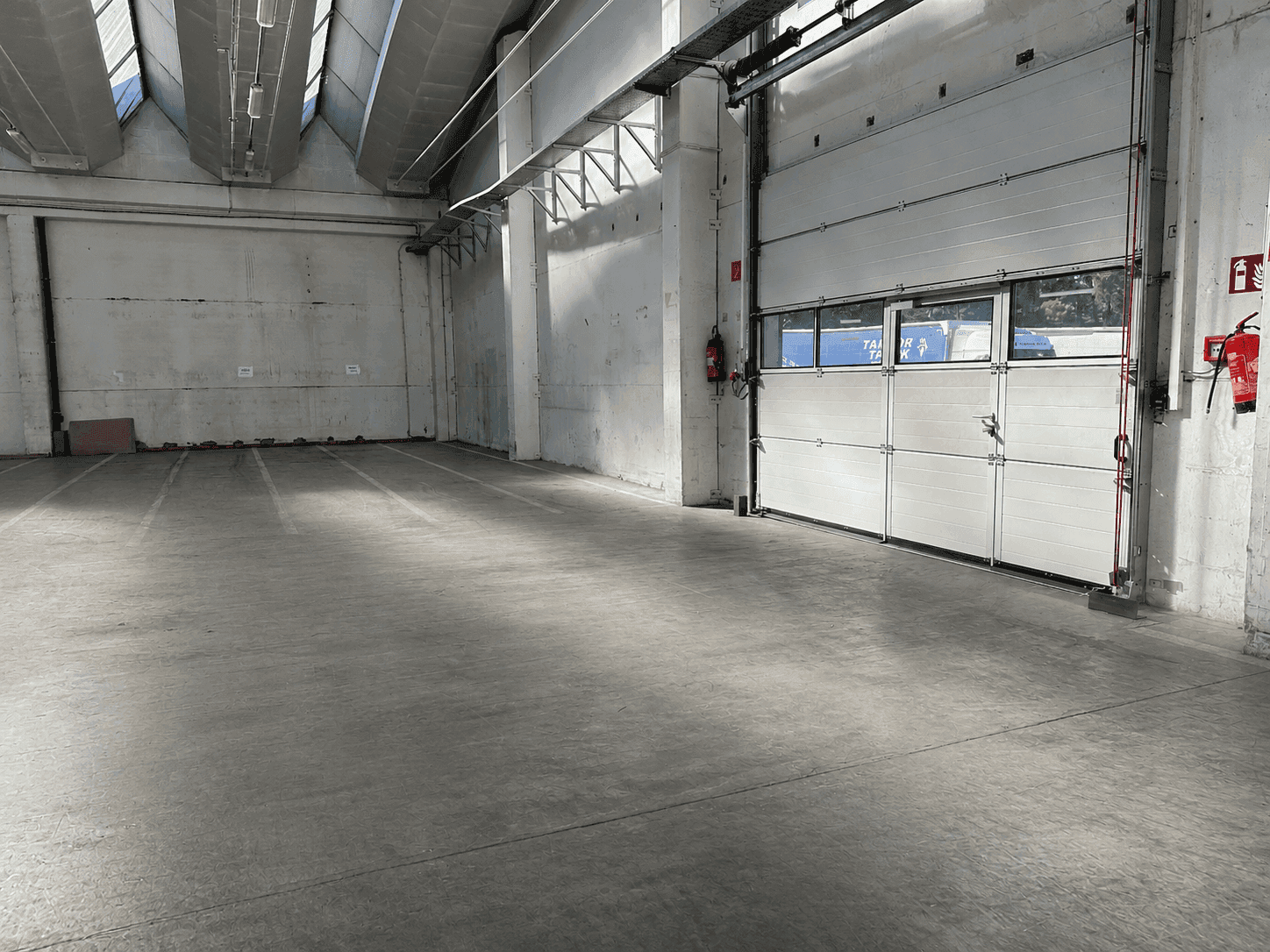 Leere Industriehalle mit Betonboden, hohen Decken und einem großen Rolltor, bereit zur Nutzung.