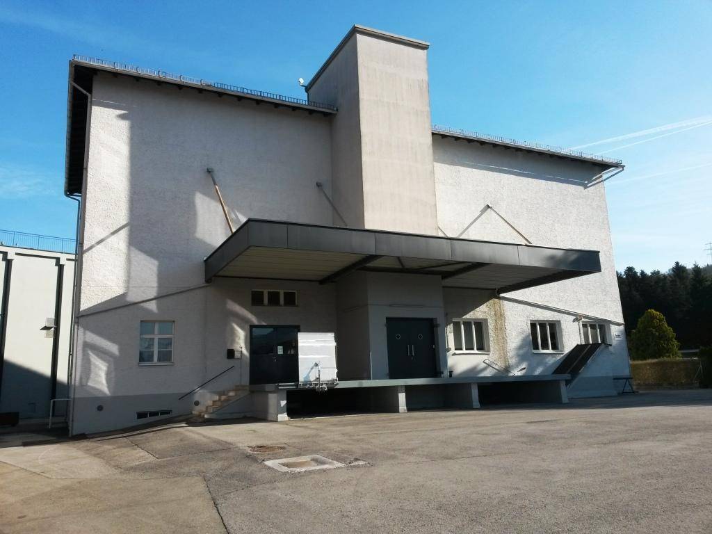 Außenansicht eines großen Industriegebäudes mit Laderampe und breiter Zufahrt unter blauem Himmel.