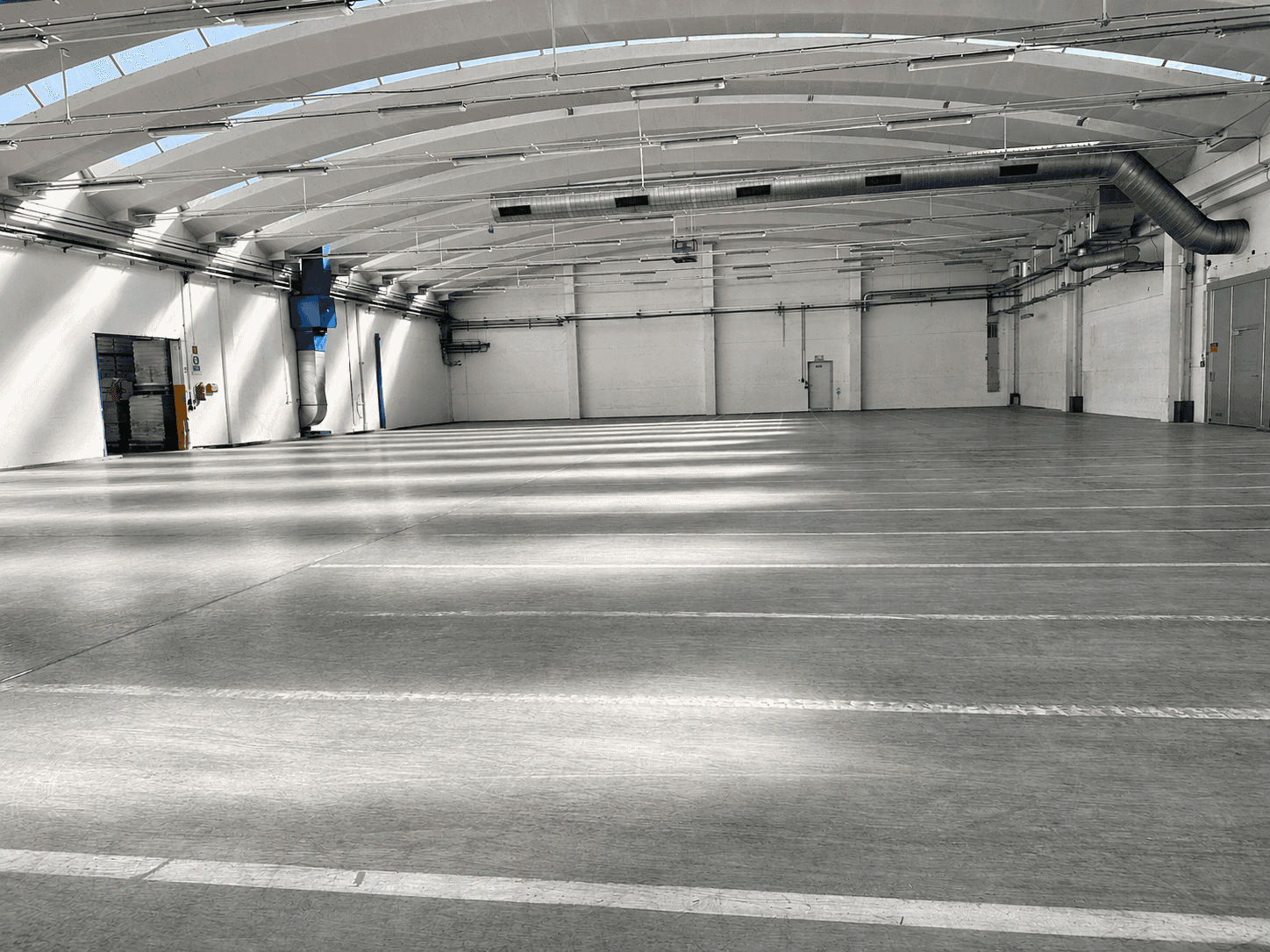 Große, leere Lagerhalle mit Betonboden, hohen Decken und Oberlichtern, die viel Tageslicht hereinlassen.