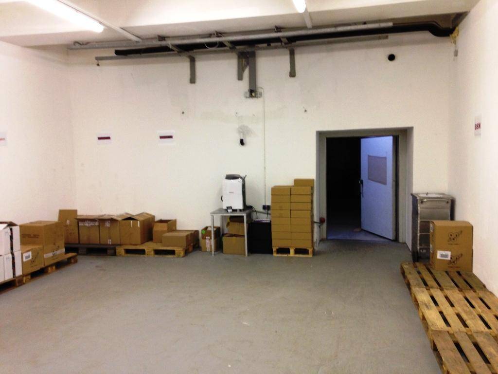 Großer Lagerraum mit offenem Durchgang, Betonboden und sichtbaren Rohrleitungen an der Decke.