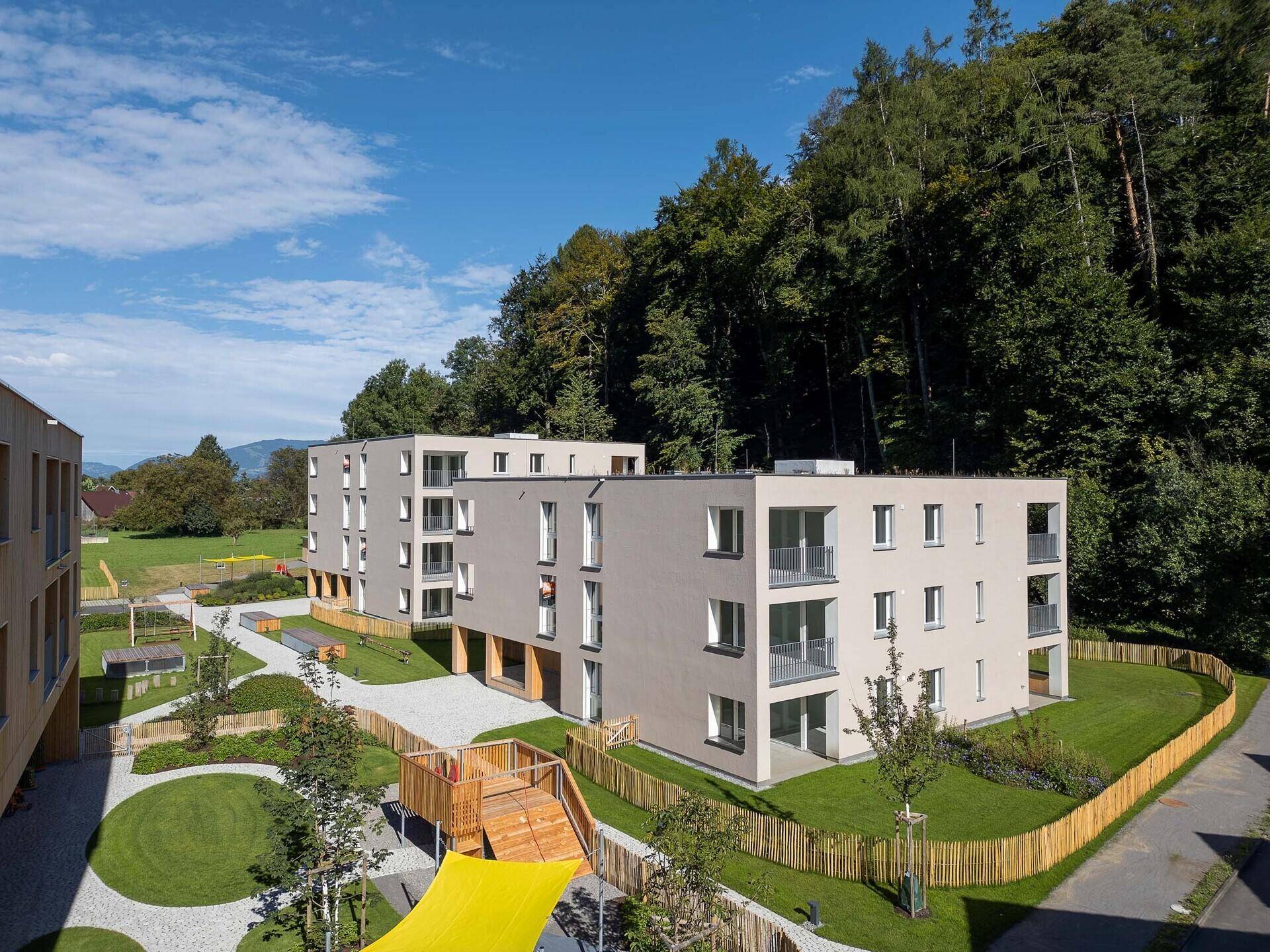 Wohnanlage mit modernen Gebäuden, Spielplatz und viel Grün in waldreicher Umgebung.