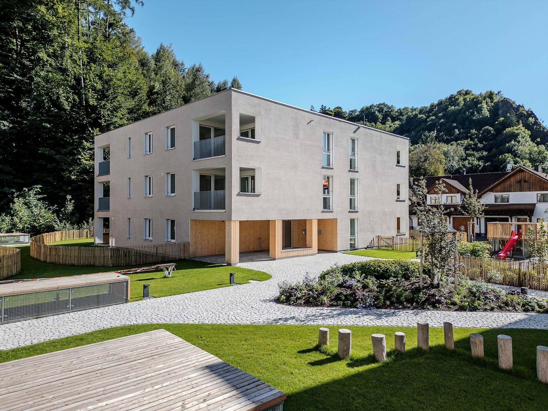 Modernes Mehrfamilienhaus mit heller Fassade, Balkonen und Spielplatz in grüner Umgebung am Waldrand.