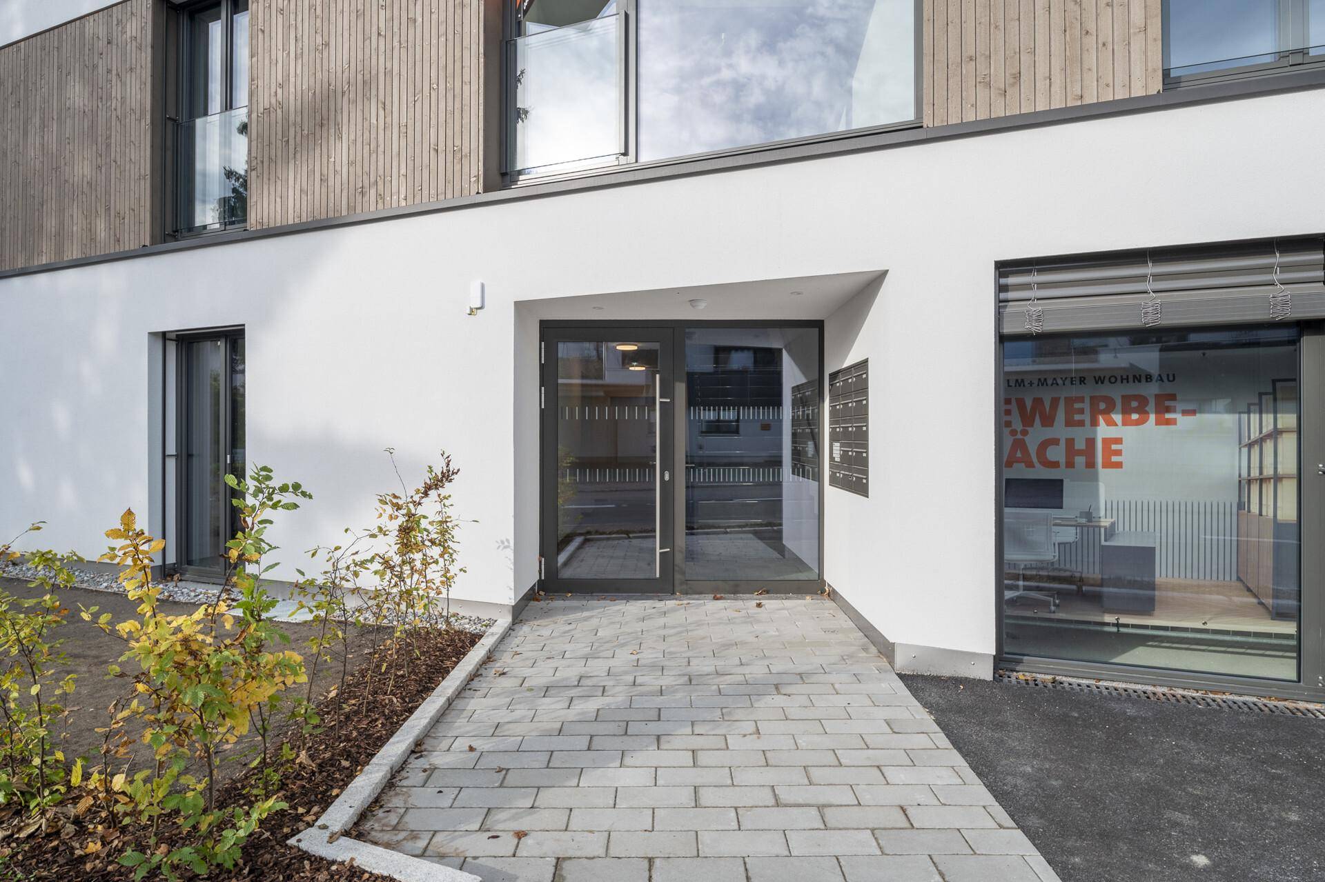 Eingangsbereich eines Neubaus mit moderner Fassade und gepflastertem Zugangsweg.