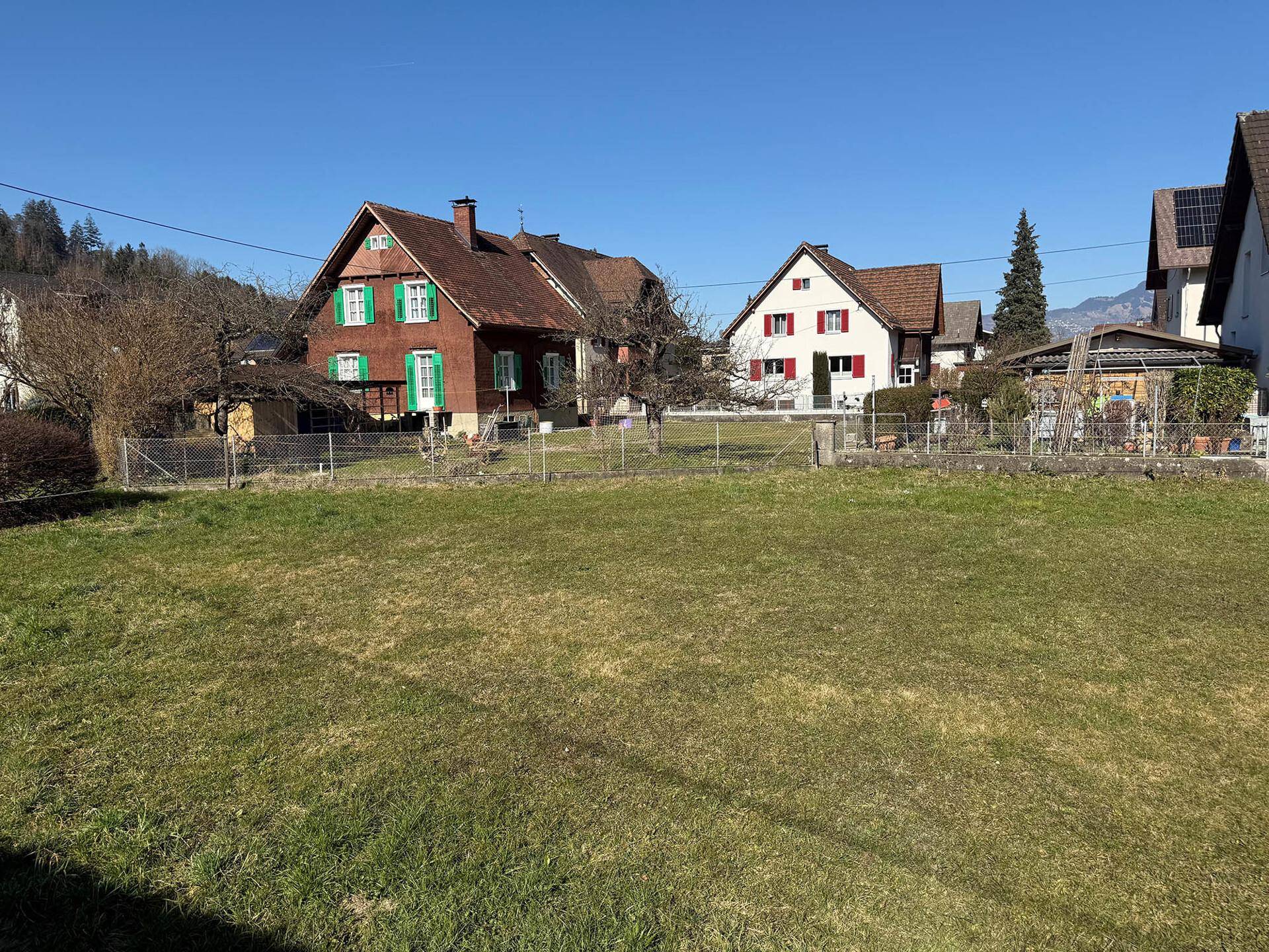 Sonniges Grundstück mit Rasenfläche und traditionellen Wohnhäusern im Hintergrund.