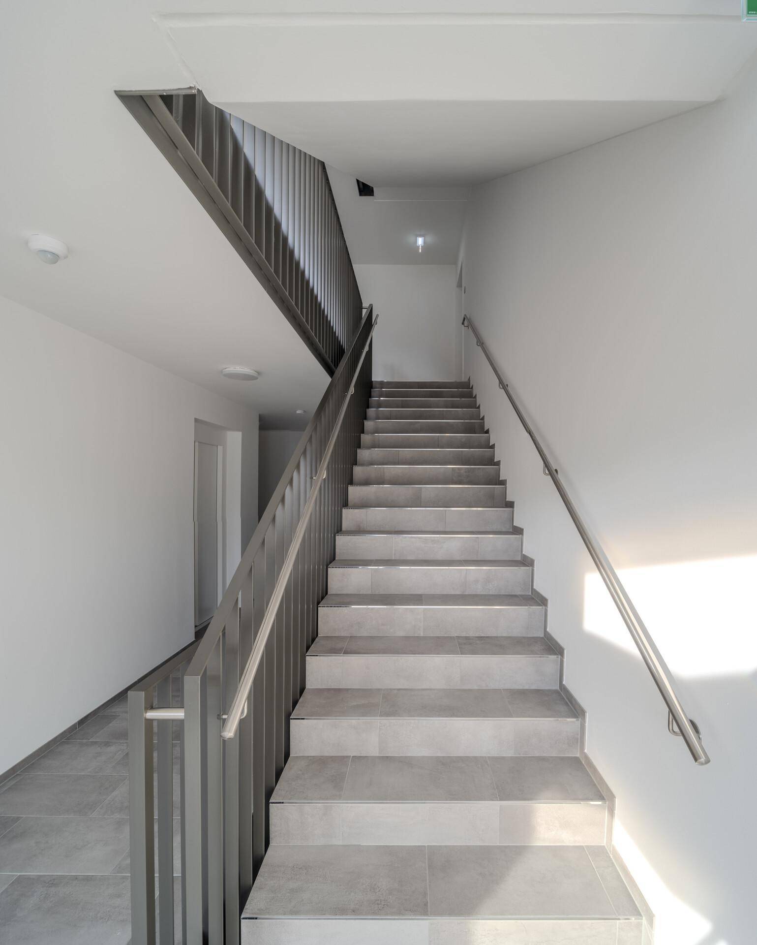 Helles Treppenhaus mit Fliesenboden und schlichtem Geländer in einem Neubau.
