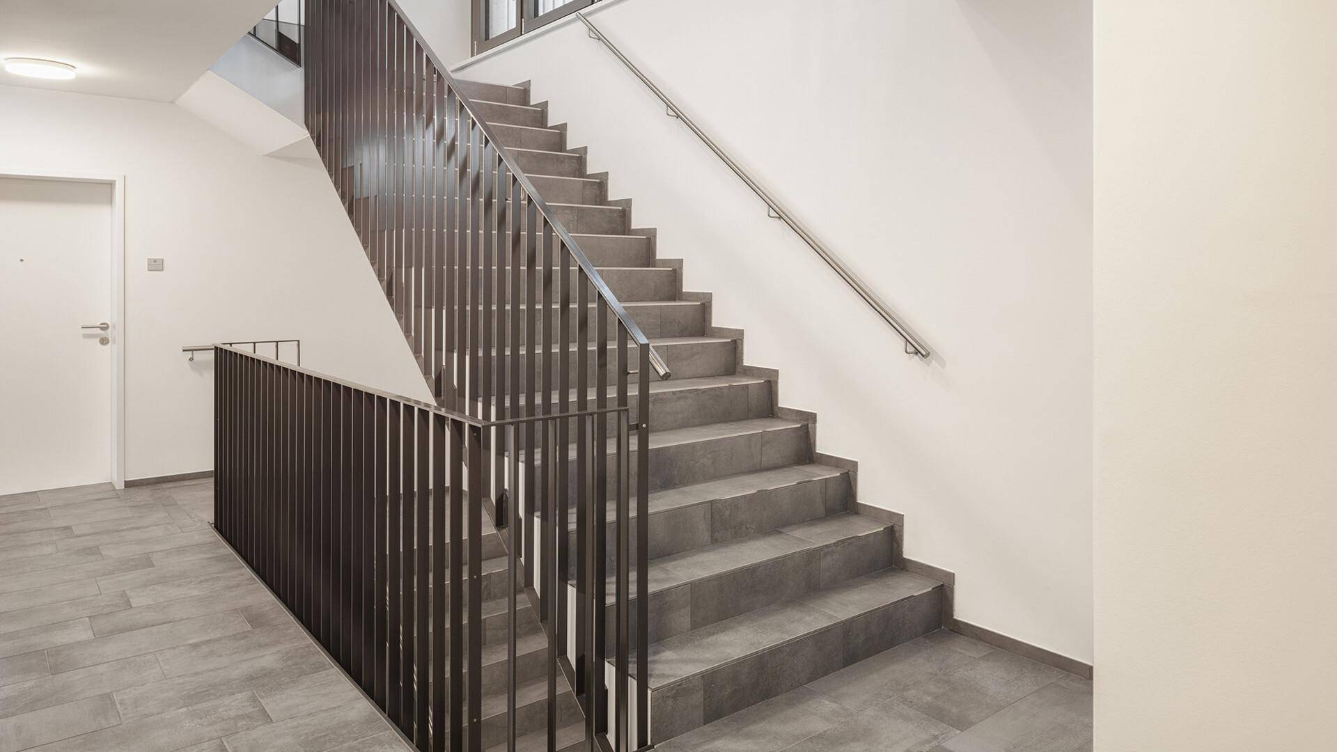 Modernes Treppenhaus mit grauen Fliesen, weißen Wänden und dunklem Metallgeländer.