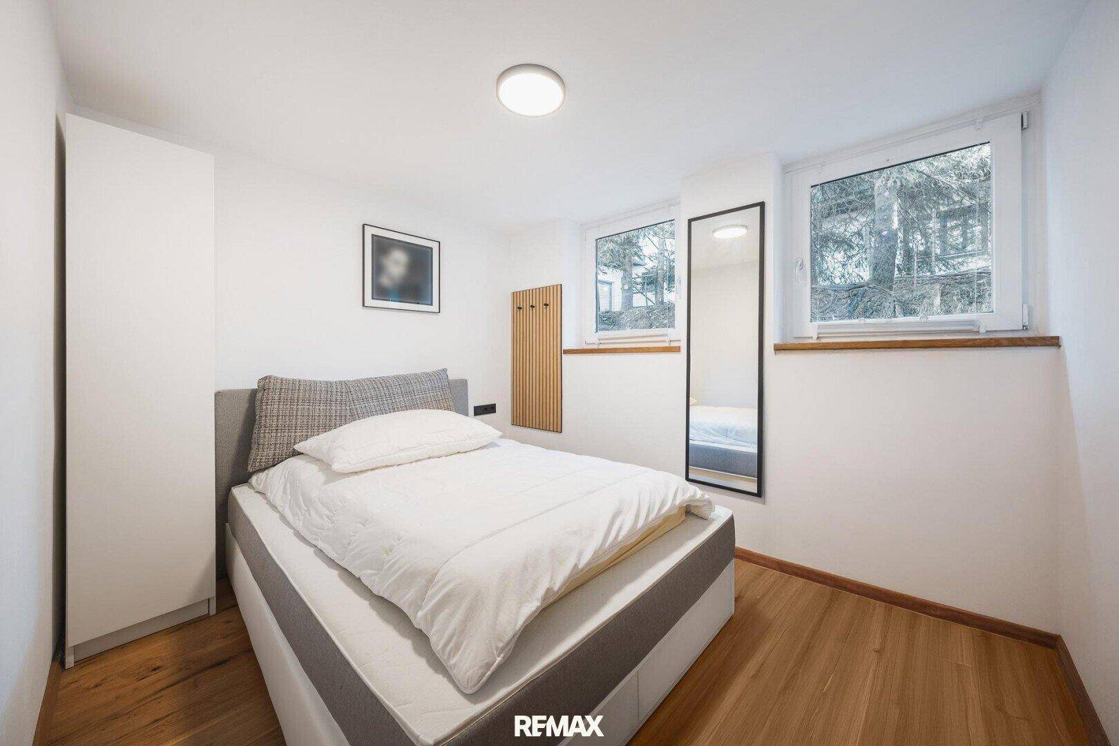 Helles Schlafzimmer mit Doppelbett, Kleiderschrank und zwei Fenstern.