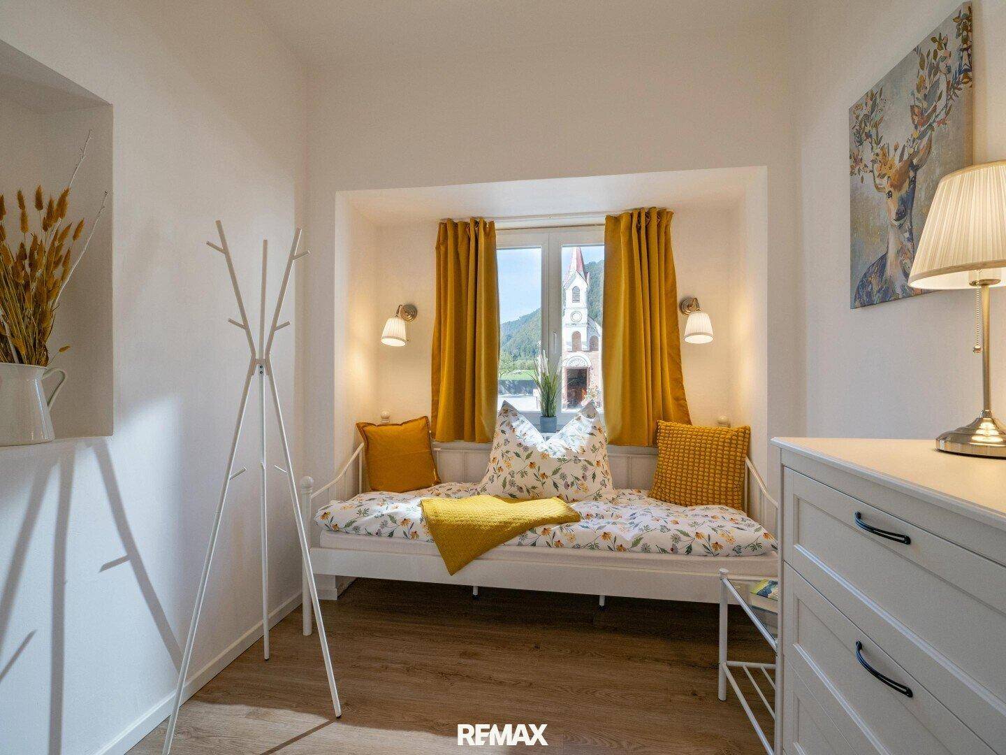 Helles Schlafzimmer mit einem Tagesbett in einer Fensternische und gelben Vorhängen, mit Bergblick.