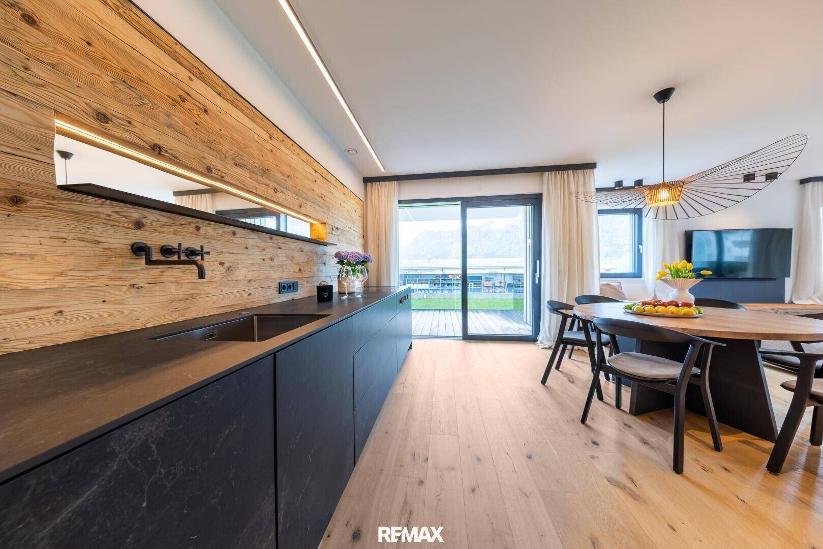 Moderne Küche mit dunklen Arbeitsplatten, Holzwand und Zugang zur Terrasse mit Bergblick.