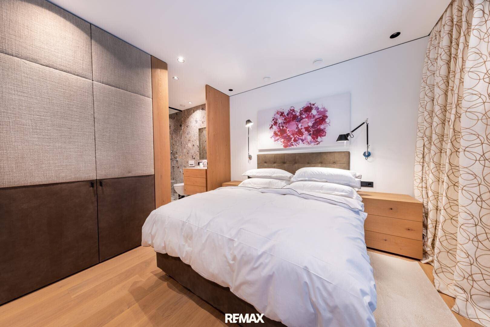 Schlafzimmer mit integrierten Kleiderschränken, Holzboden und Zugang zu einem modernen Badezimmer.
