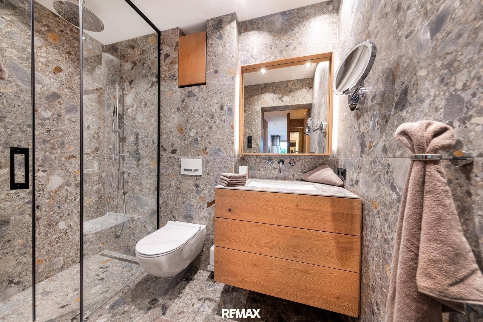 Modernes Badezimmer mit begehbarer Dusche, Wand-WC und Holzwaschtisch. Die Steinoptik verleiht eine natürliche Note.