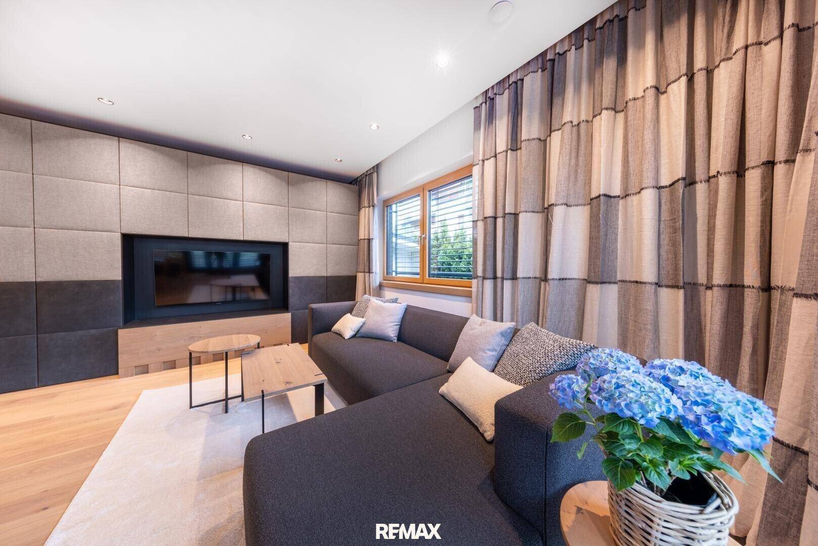 Gemütliches Wohnzimmer mit modernem Kamin, bequemer Couch und Holzboden. Die Fenster bieten einen Blick ins Grüne.