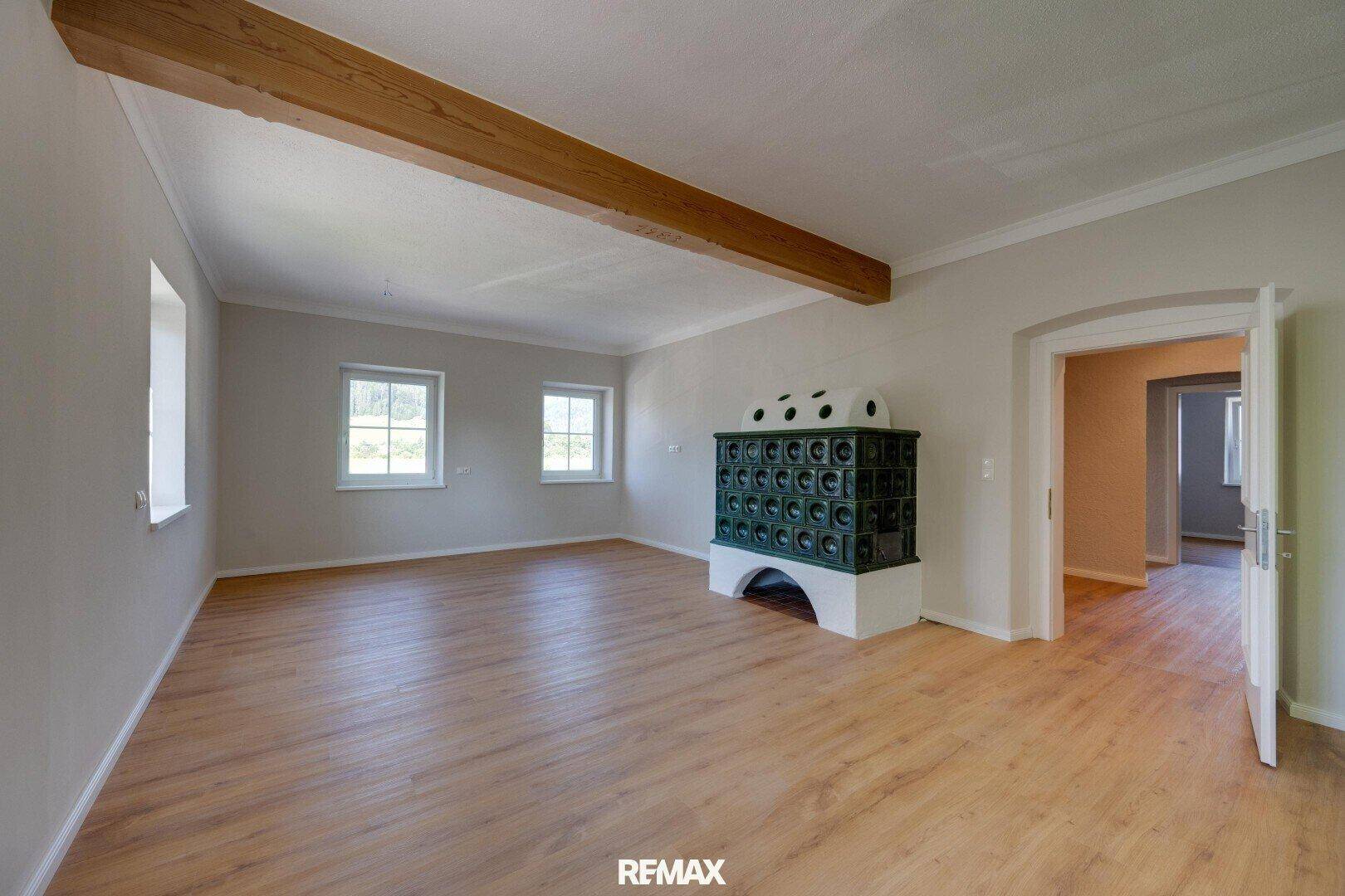 Geräumiges Wohnzimmer mit Holzboden, einem traditionellen Kachelofen und sichtbarem Deckenbalken.