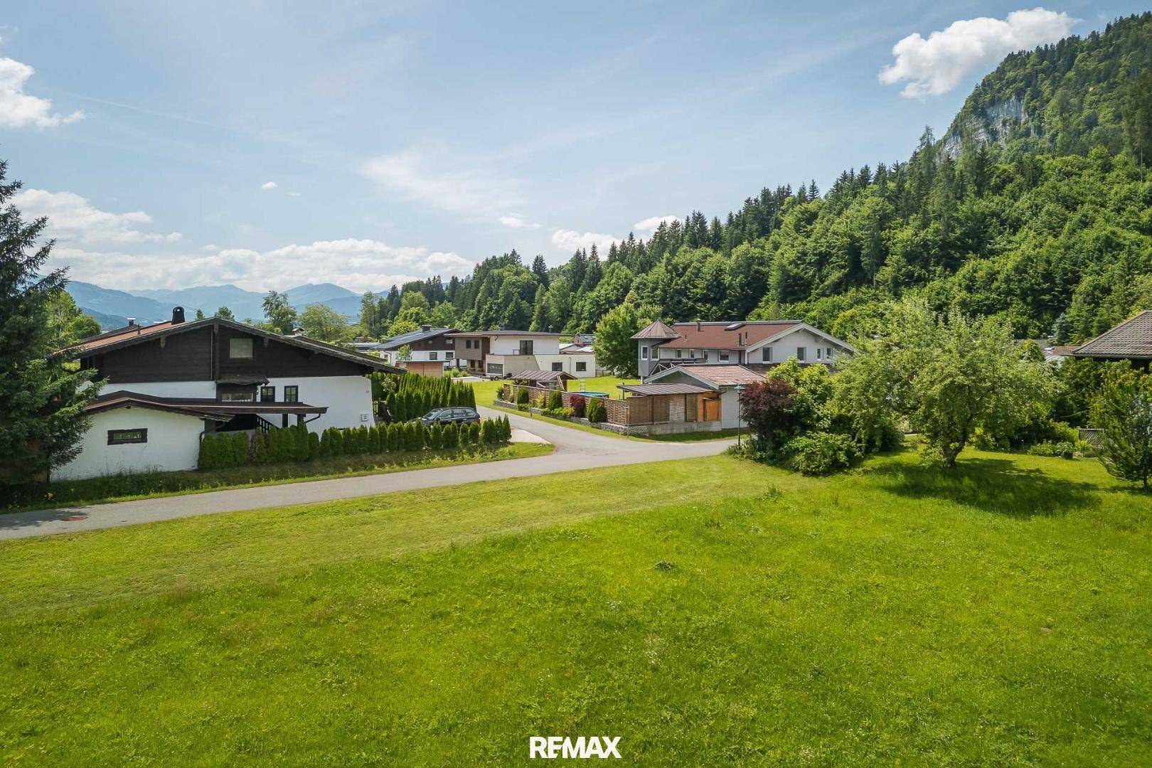 Grüne Wiesen und Wohnhäuser am Fuße eines bewaldeten Berges unter blauem Himmel.