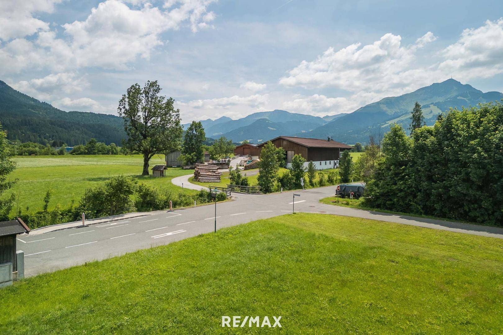 Ländliche Straße führt durch grüne Felder mit einem Bauernhof und Bergen am Horizont.
