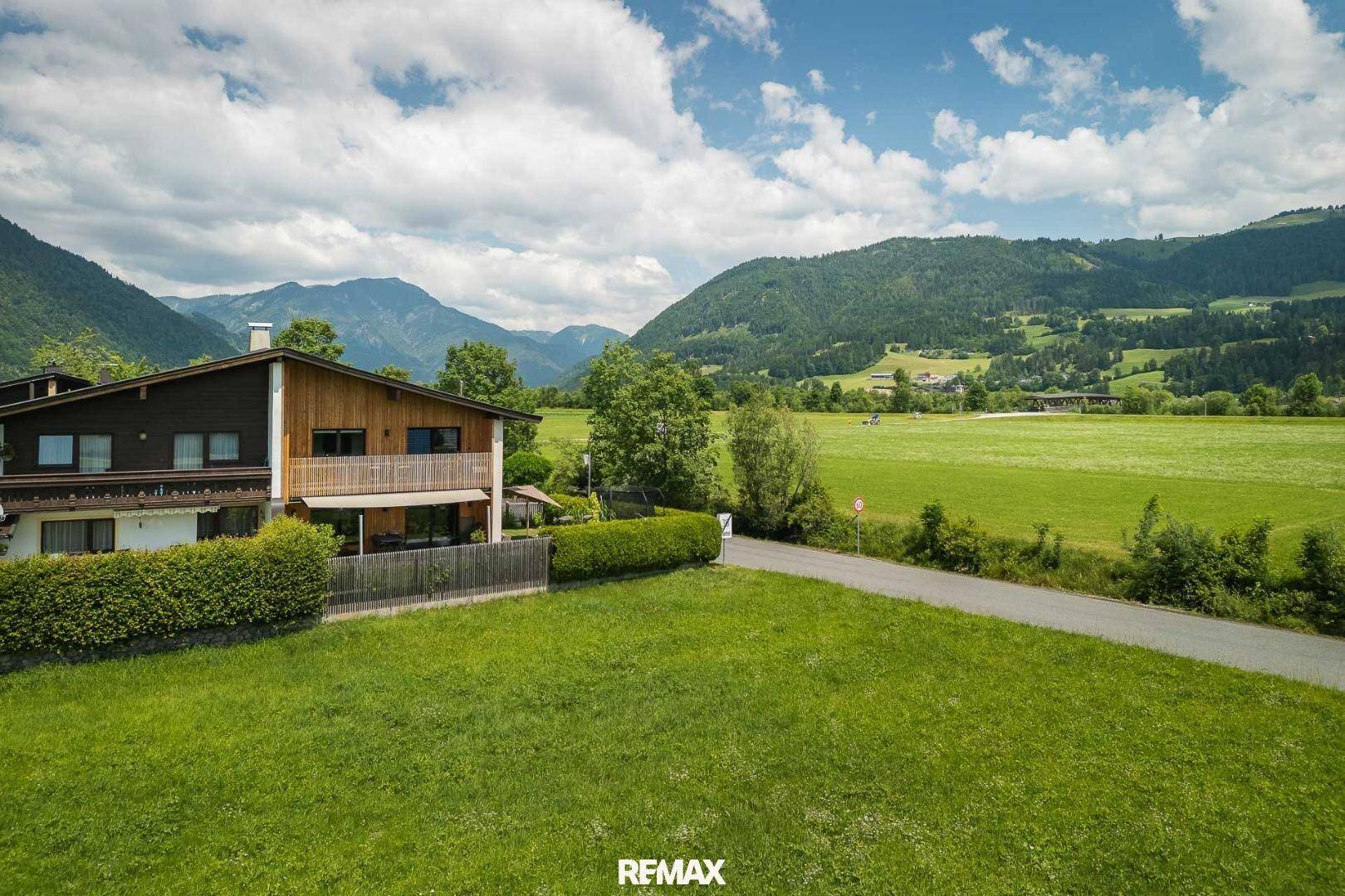 Hausfassade mit Balkon, umgeben von einer gepflegten Hecke, Rasenflächen und einer malerischen Berglandschaft.