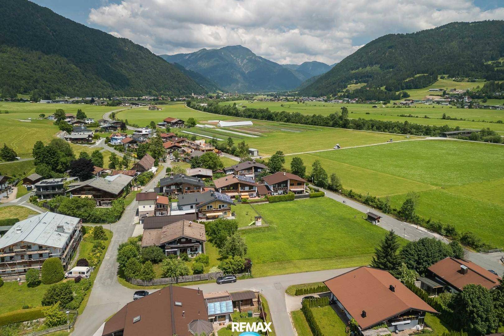 Panoramablick aus der Luft auf eine ländliche Wohngegend, umgeben von weiten Feldern und einer beeindruckenden Bergkulisse.