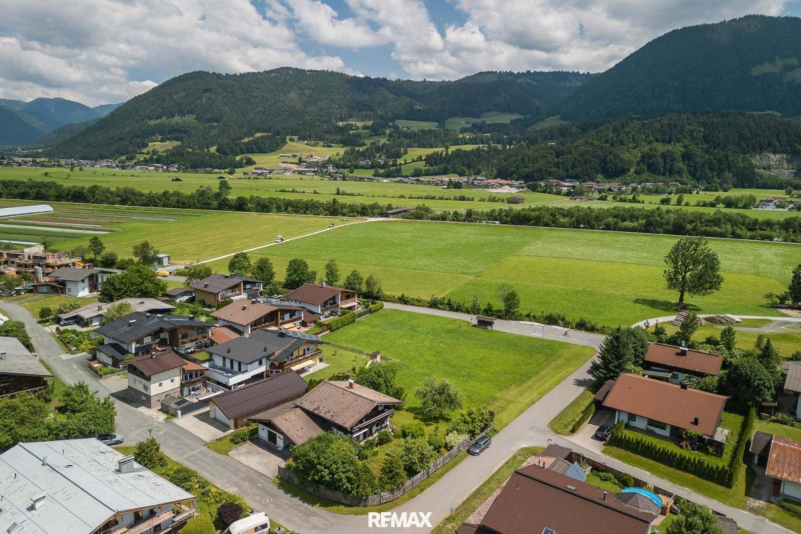 Weitläufige Luftaufnahme einer Wohnsiedlung mit grünen Feldern und majestätischen Bergen im Hintergrund.