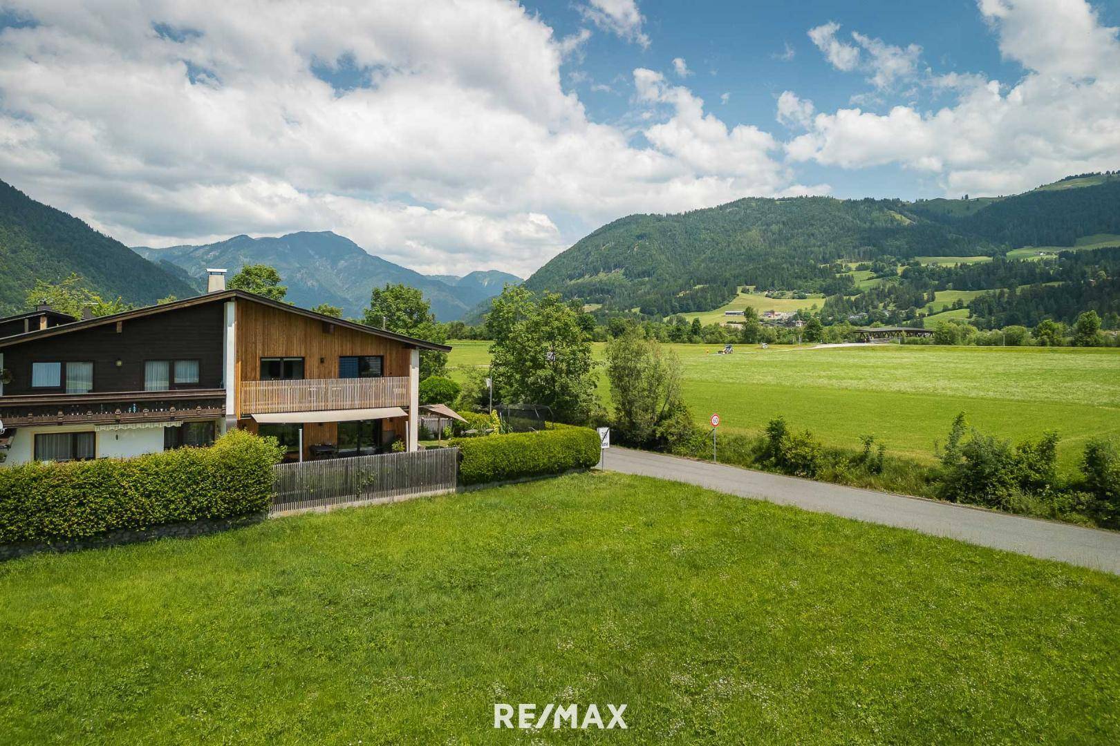 Haus mit Holzbalkon und gepflegtem Rasen, umgeben von weiten Feldern und Bergen.