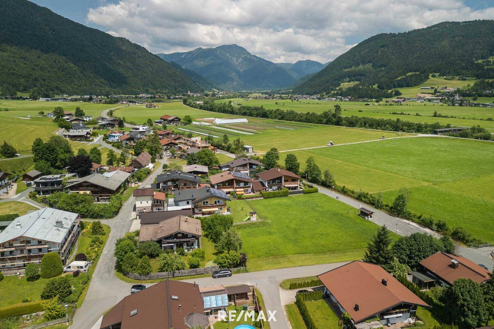 Weitläufige Ansicht einer Wohnsiedlung in einem Tal, mit Feldern und einer beeindruckenden Bergkulisse.