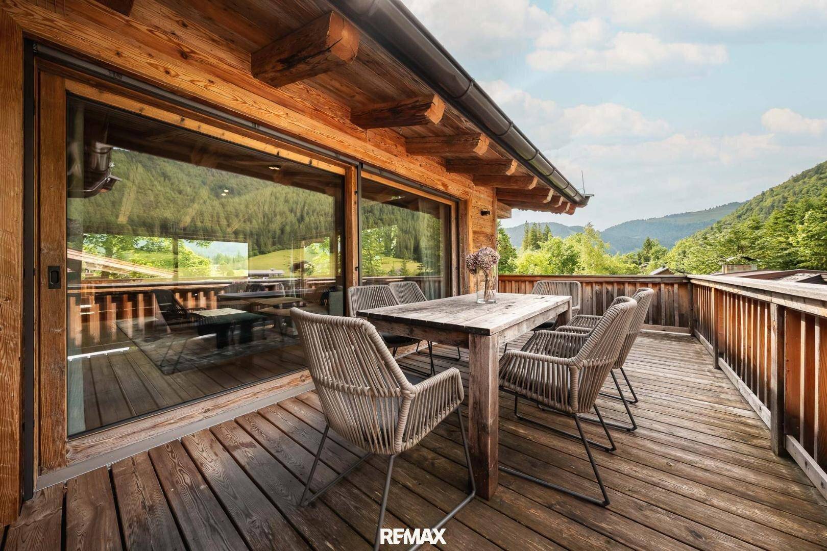Geräumige Holzterrasse mit Esstisch und Stühlen, umgeben von grünen Bergen unter blauem Himmel.