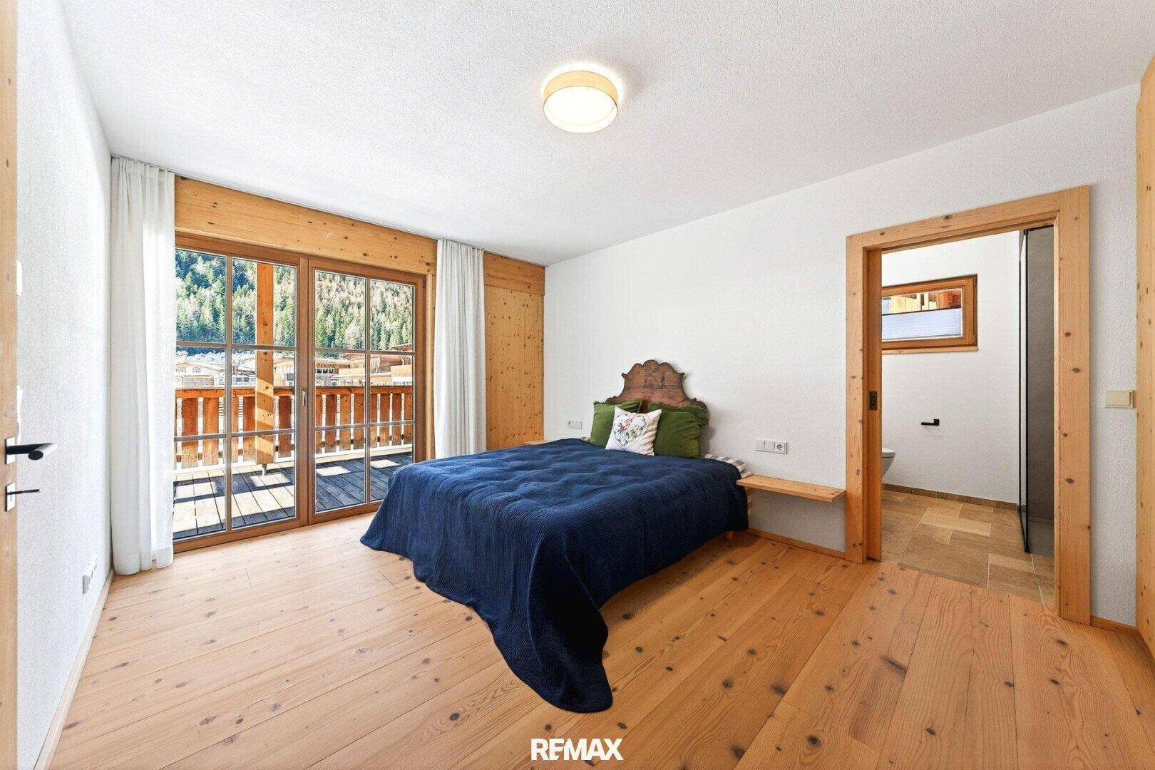 Helles Schlafzimmer mit Holzboden, Zugang zum Balkon und Blick auf die umliegende Berglandschaft.