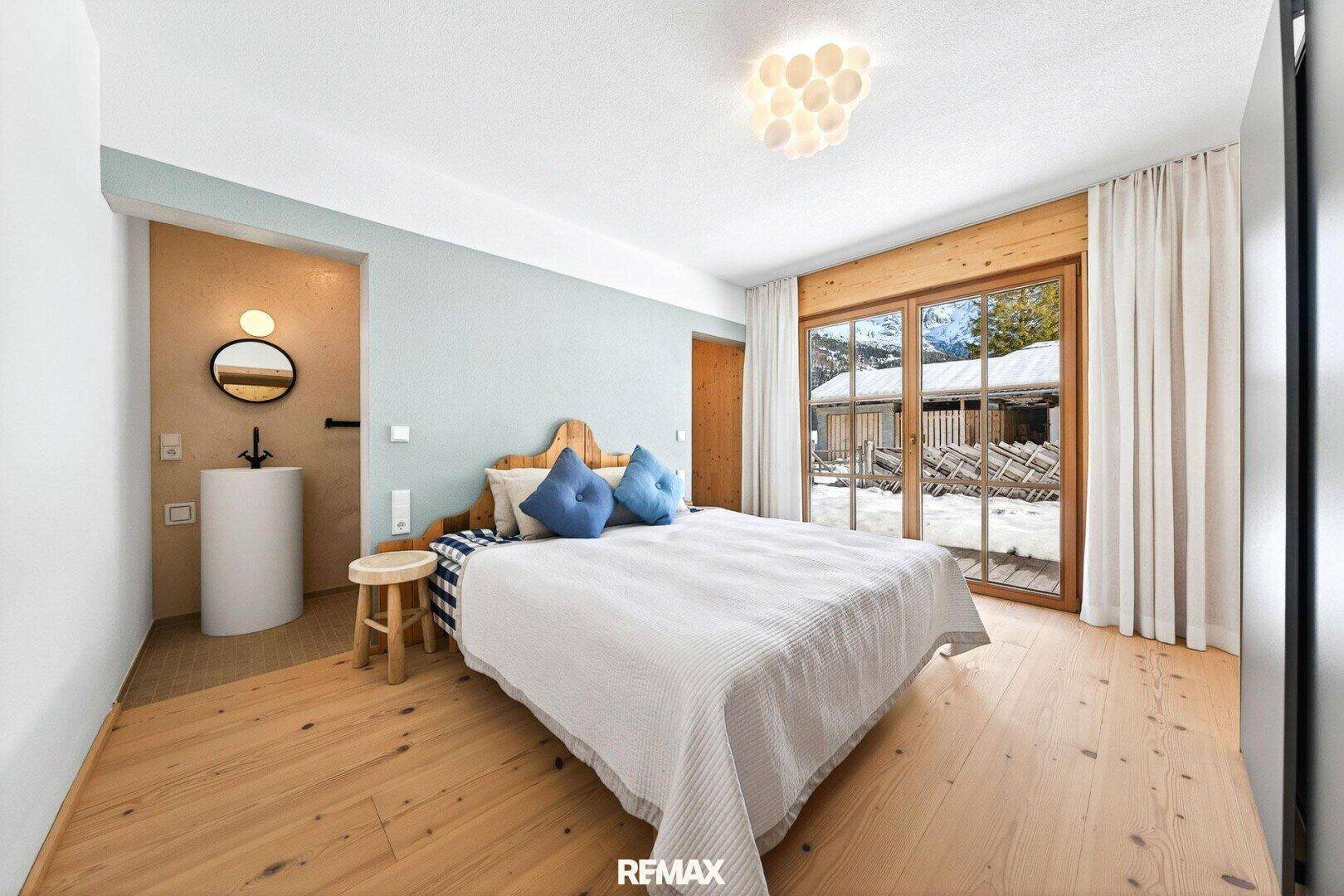 Gemütliches Schlafzimmer mit Holzboden, großem Fenster und integriertem Waschbeckenbereich.