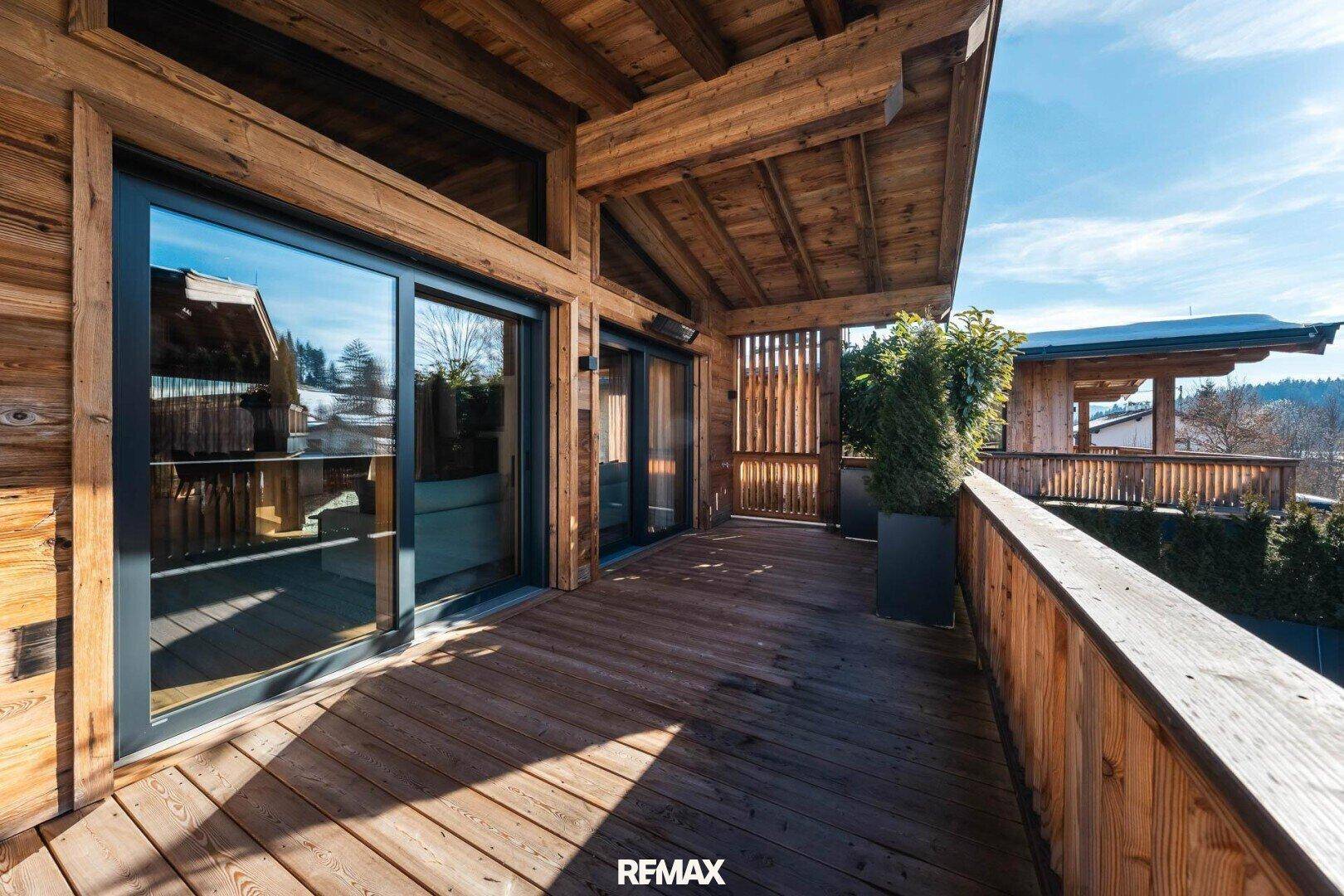 Geräumiger Holzbalkon mit großen Glasfronten und Blick auf die grüne Umgebung.