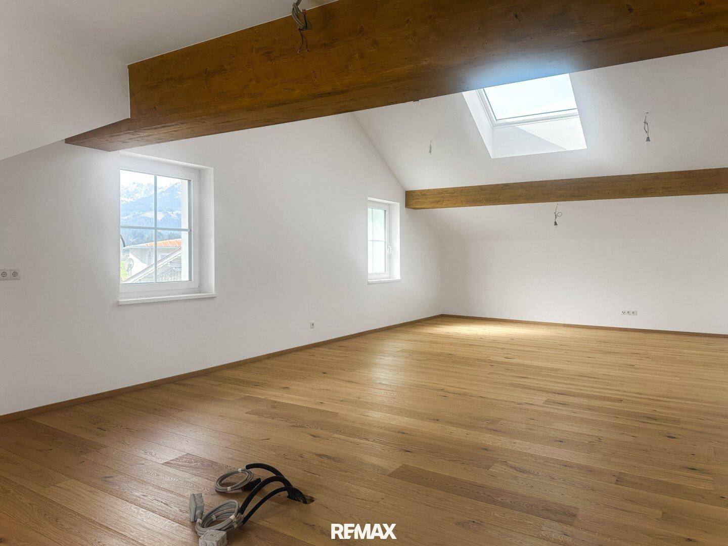 Heller, leerer Raum mit Holzbalken, schrägen Decken, Holzboden und Dachfenster.