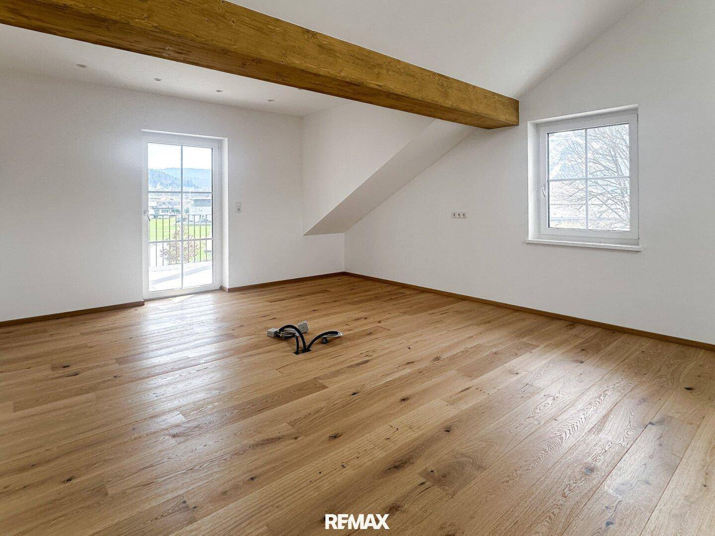 Geräumiger, leerer Raum mit Holzboden, Holzbalken und Zugang zu einem Balkon oder einer Terrasse.