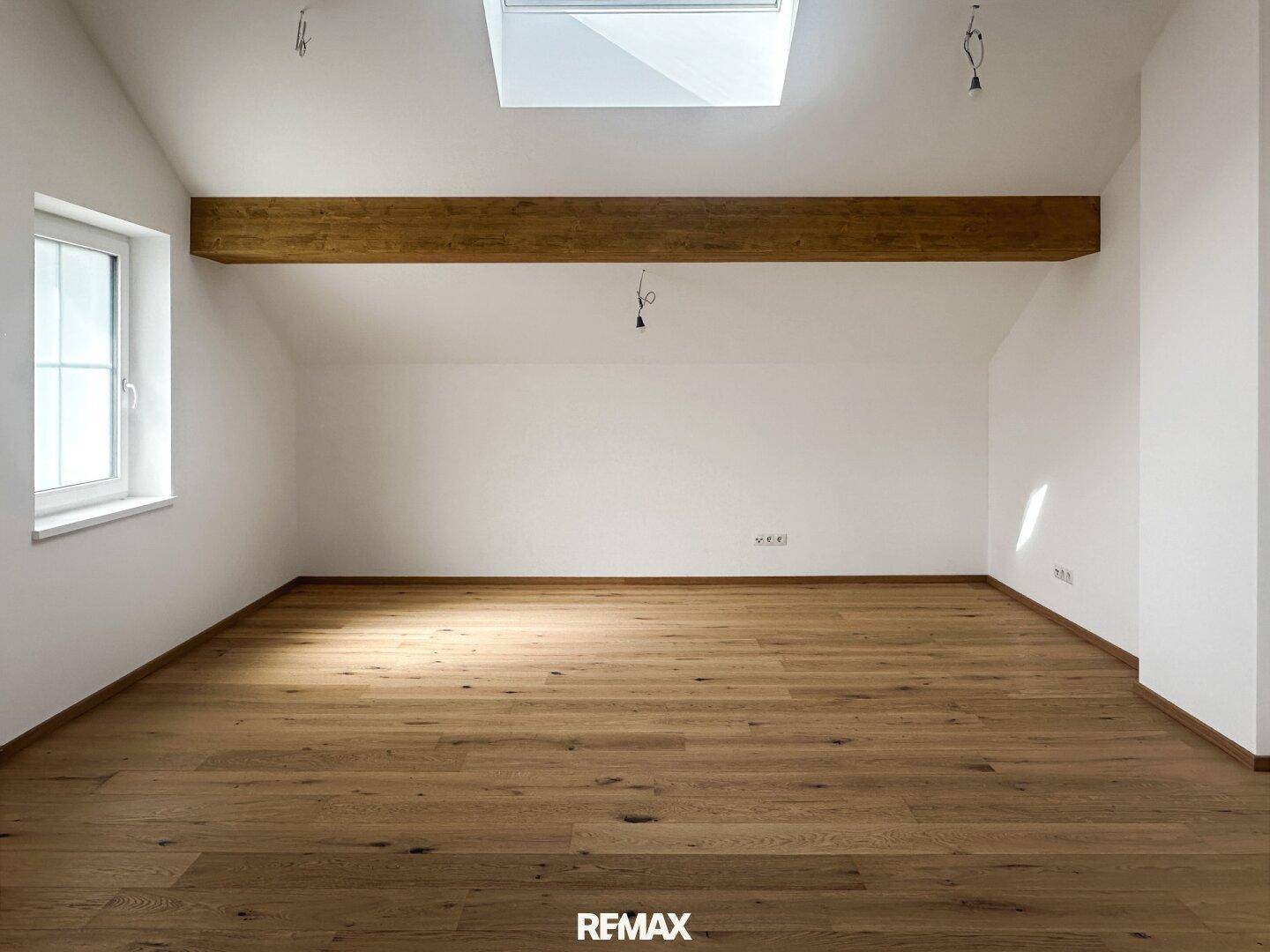 Heller, leerer Raum mit Holzboden, Holzbalken und einem Dachfenster, das viel Licht spendet.