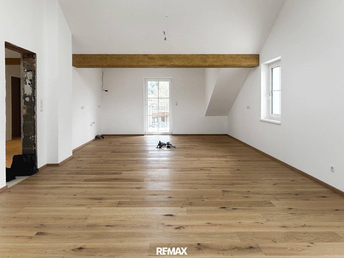 Geräumiger, leerer Raum mit Holzbalken, schrägen Decken und Zugang zu einem Außenbereich.