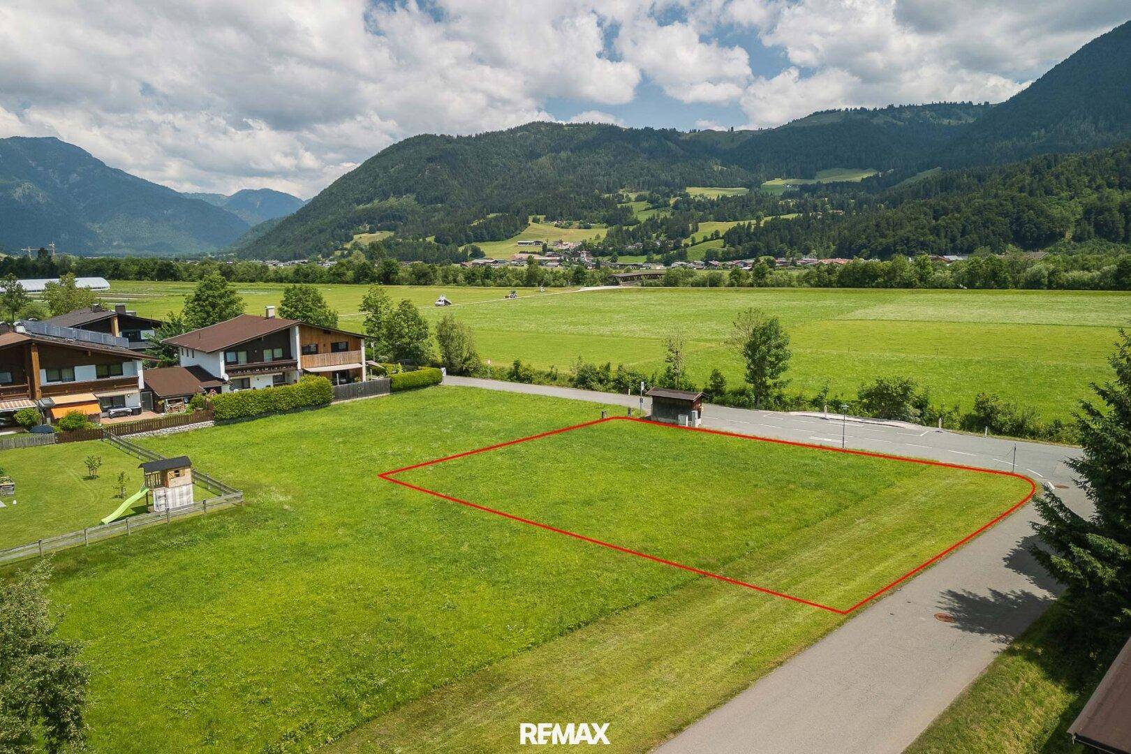 Grünes Baugrundstück mit roter Umrandung, umgeben von Natur und Berglandschaft.