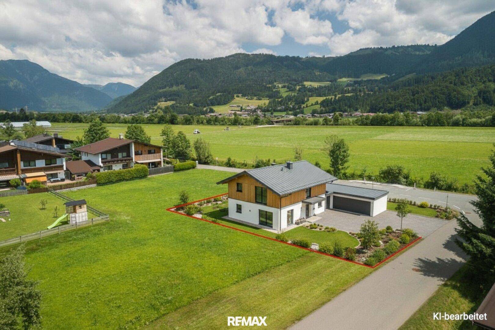 Luftaufnahme eines modernen Einfamilienhauses mit Garten, Garage und Spielplatz, umgeben von grünen Feldern und Bergen.