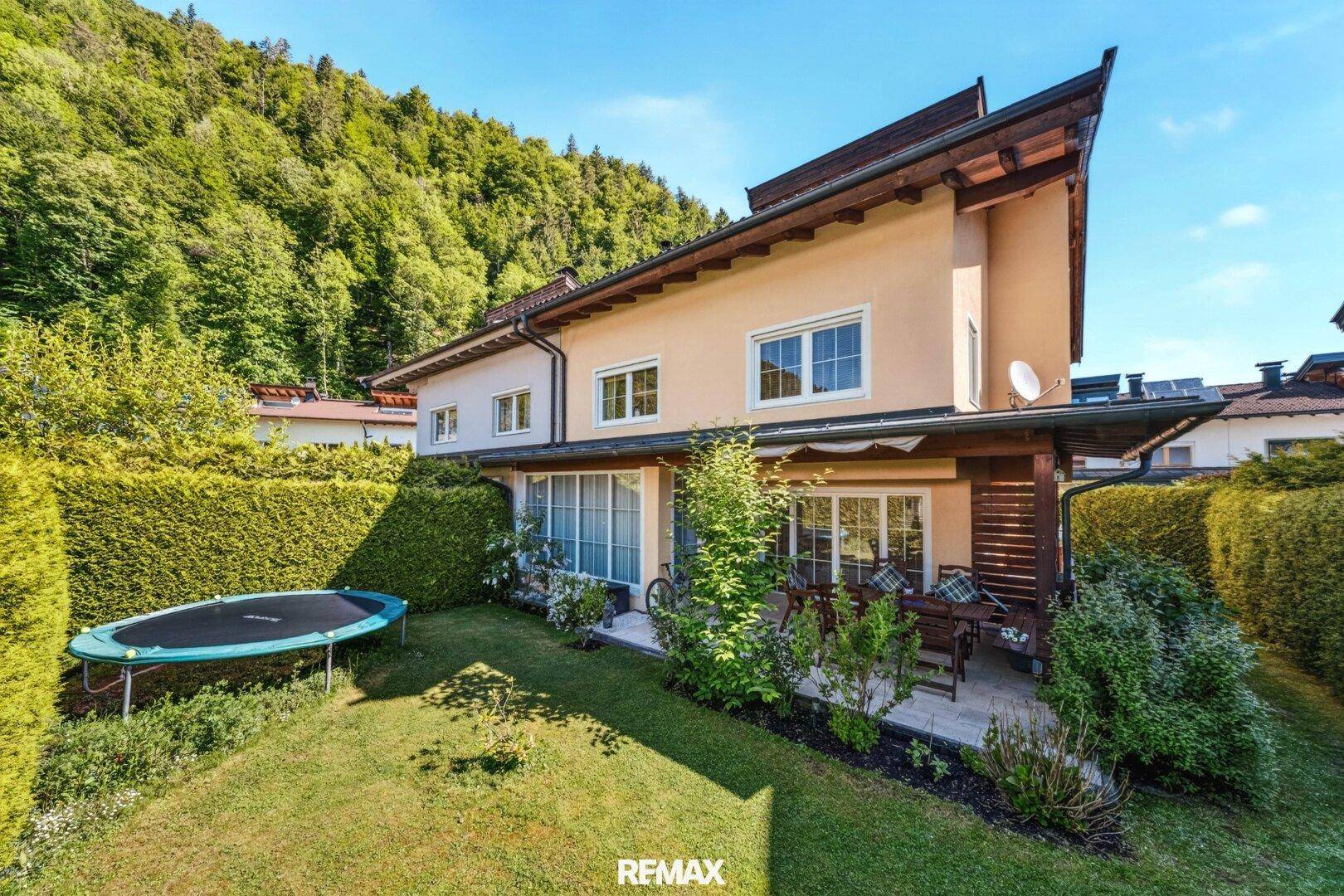 Hausfassade mit großem Garten, Trampolin und üppiger grüner Berglandschaft im Hintergrund.
