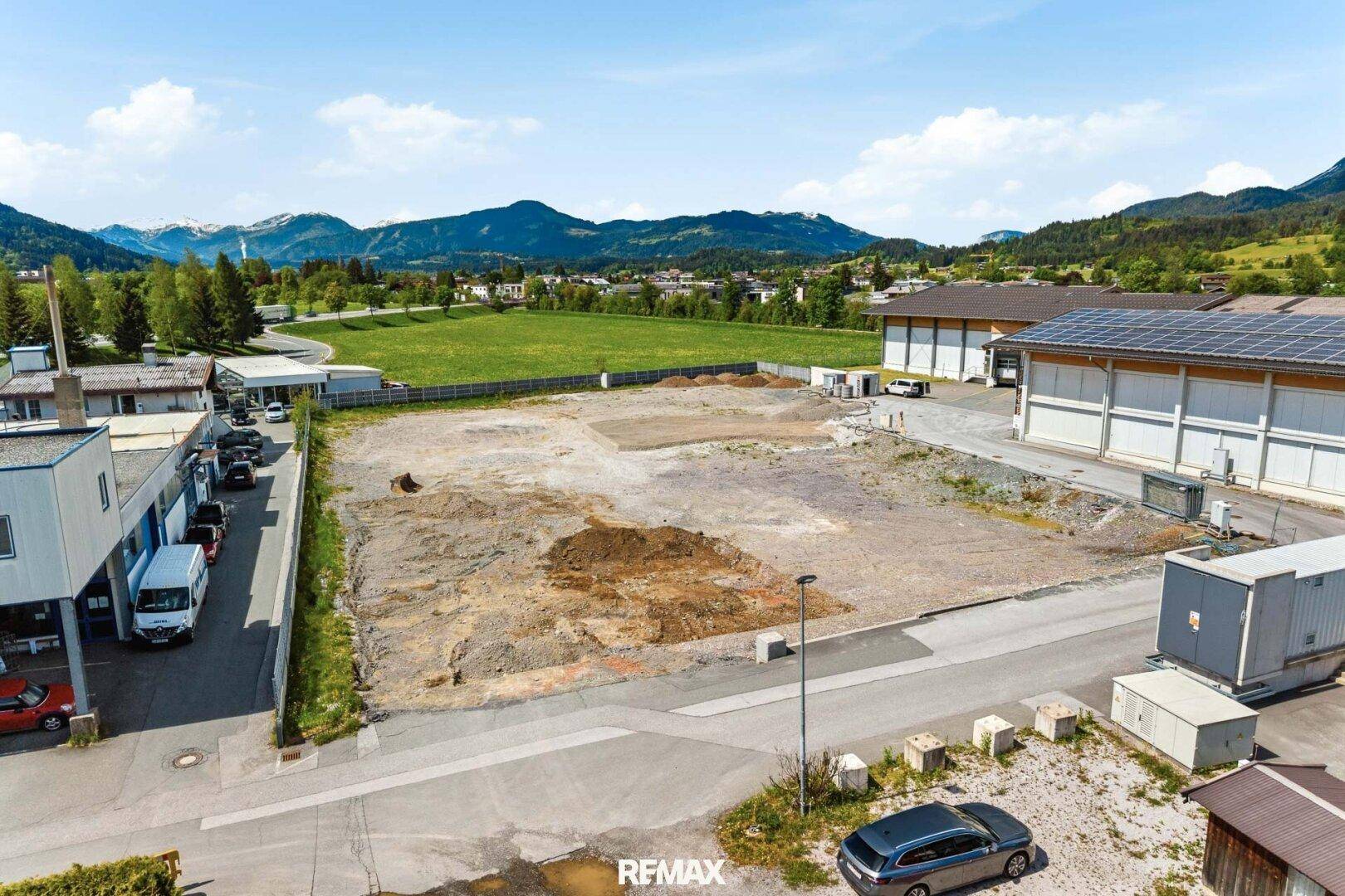Luftaufnahme eines unbebauten Grundstücks in einem Gewerbegebiet mit Bergen und umliegenden Gebäuden im Hintergrund.