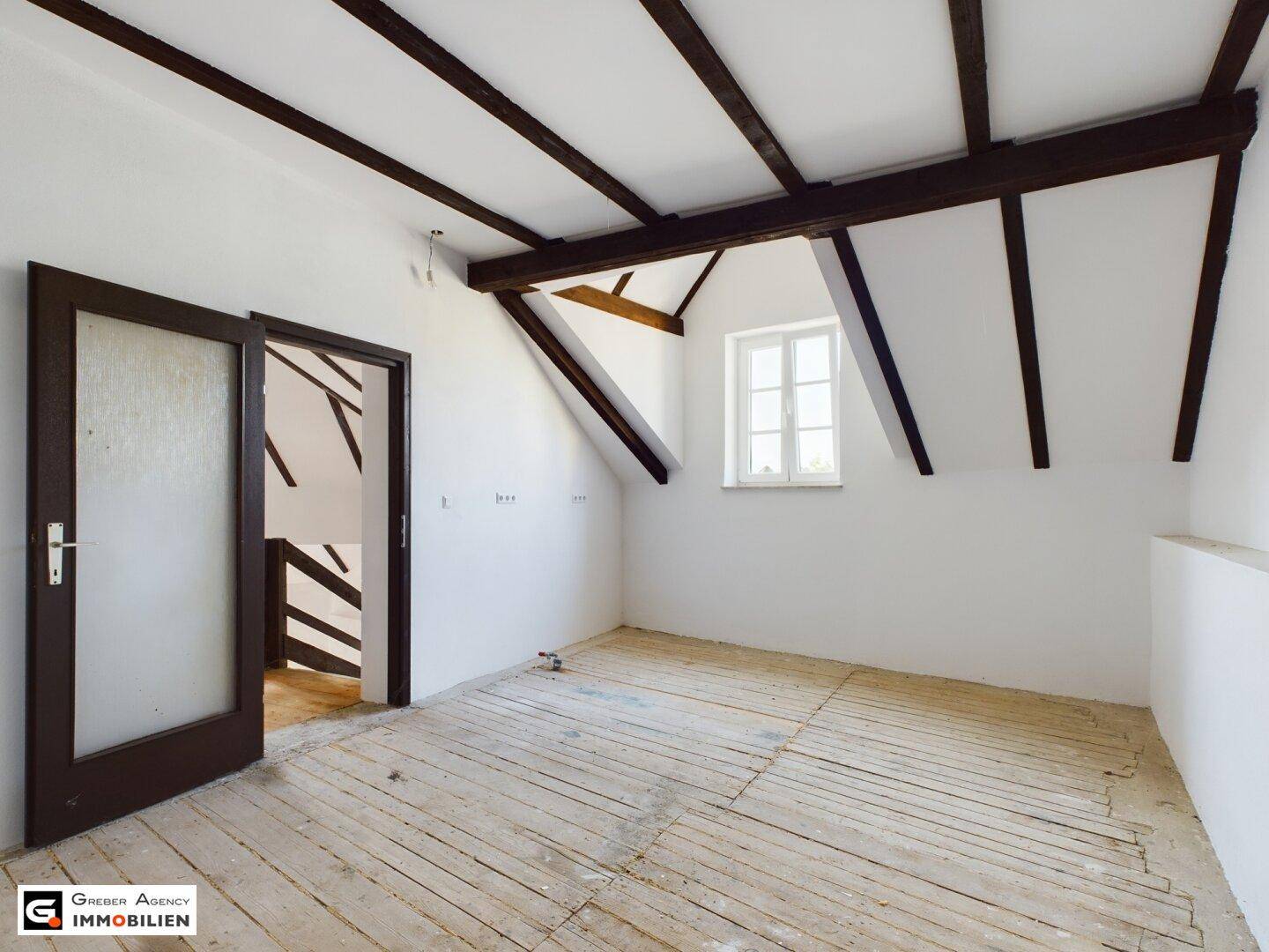 Helles Zimmer im Dachgeschoss mit schrägen Decken, sichtbaren Holzbalken und Holzdielenboden.
