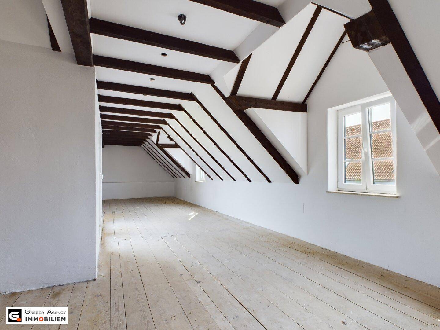 Geräumiges Dachzimmer mit freiliegenden Holzbalken, schrägen Wänden und hellem Holzdielenboden.