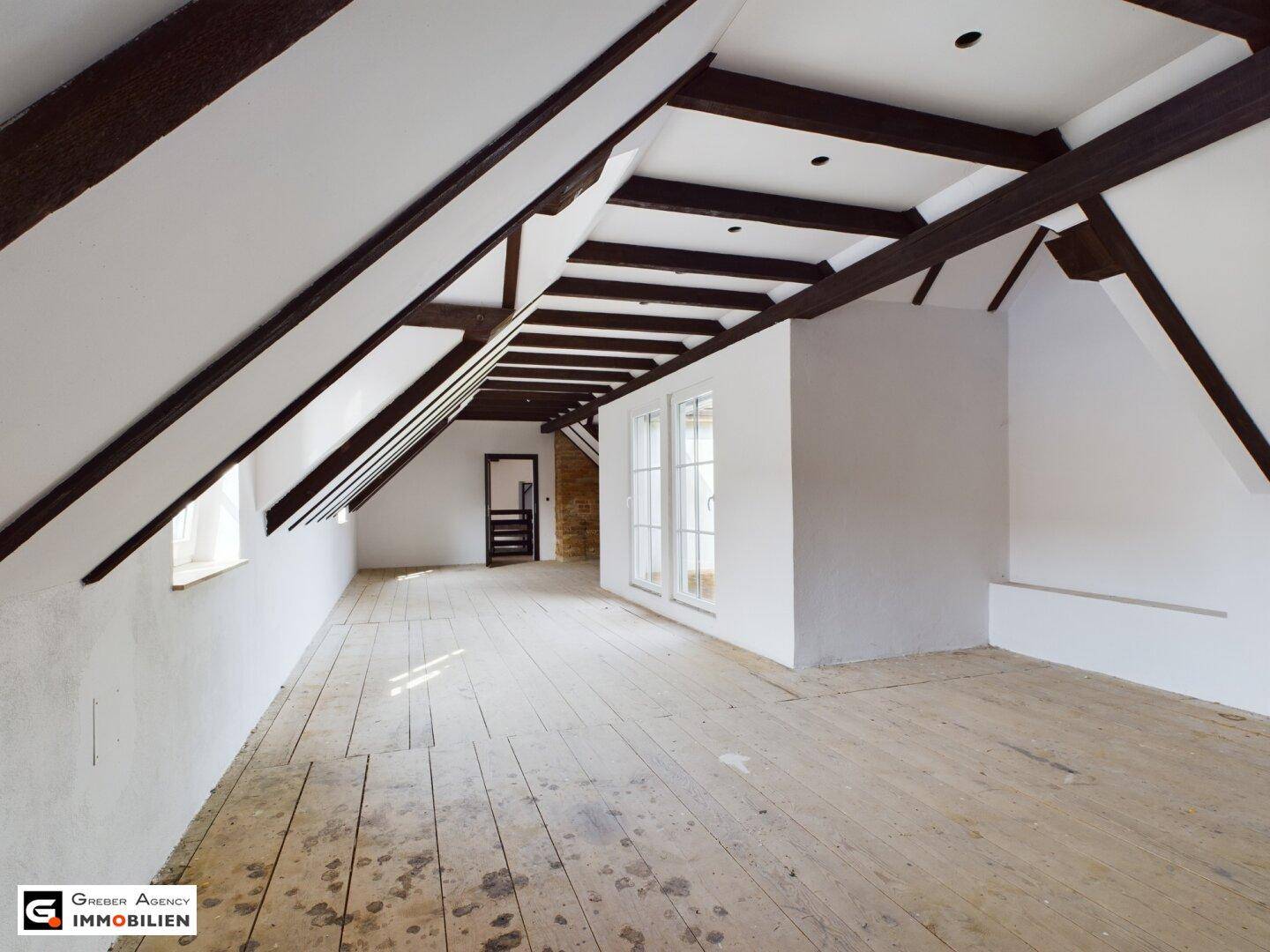 Langes Dachzimmer mit sichtbaren Holzbalken, schrägen Decken und hellem Holzdielenboden.