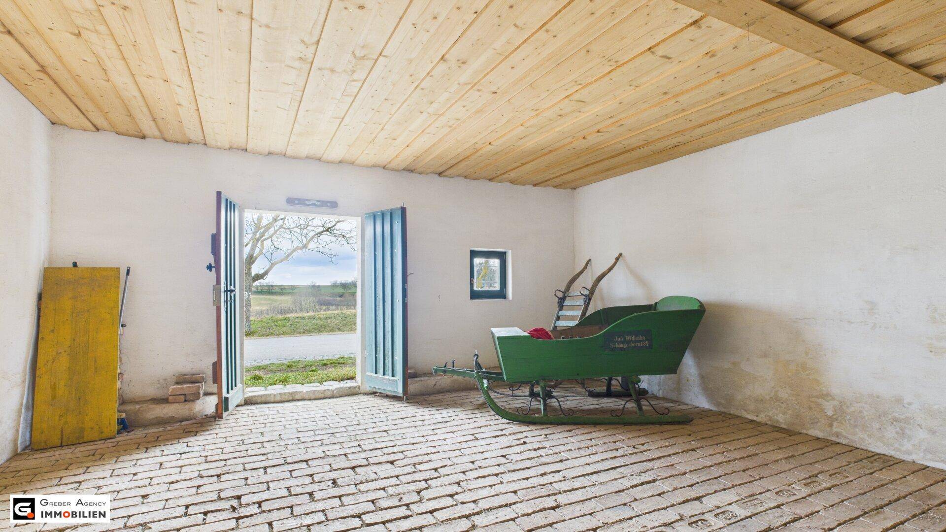 Heller Innenraum des Kellerhauses mit Holzbalkendecke, Ziegelboden und Blick nach draußen durch die offene grüne Tür.