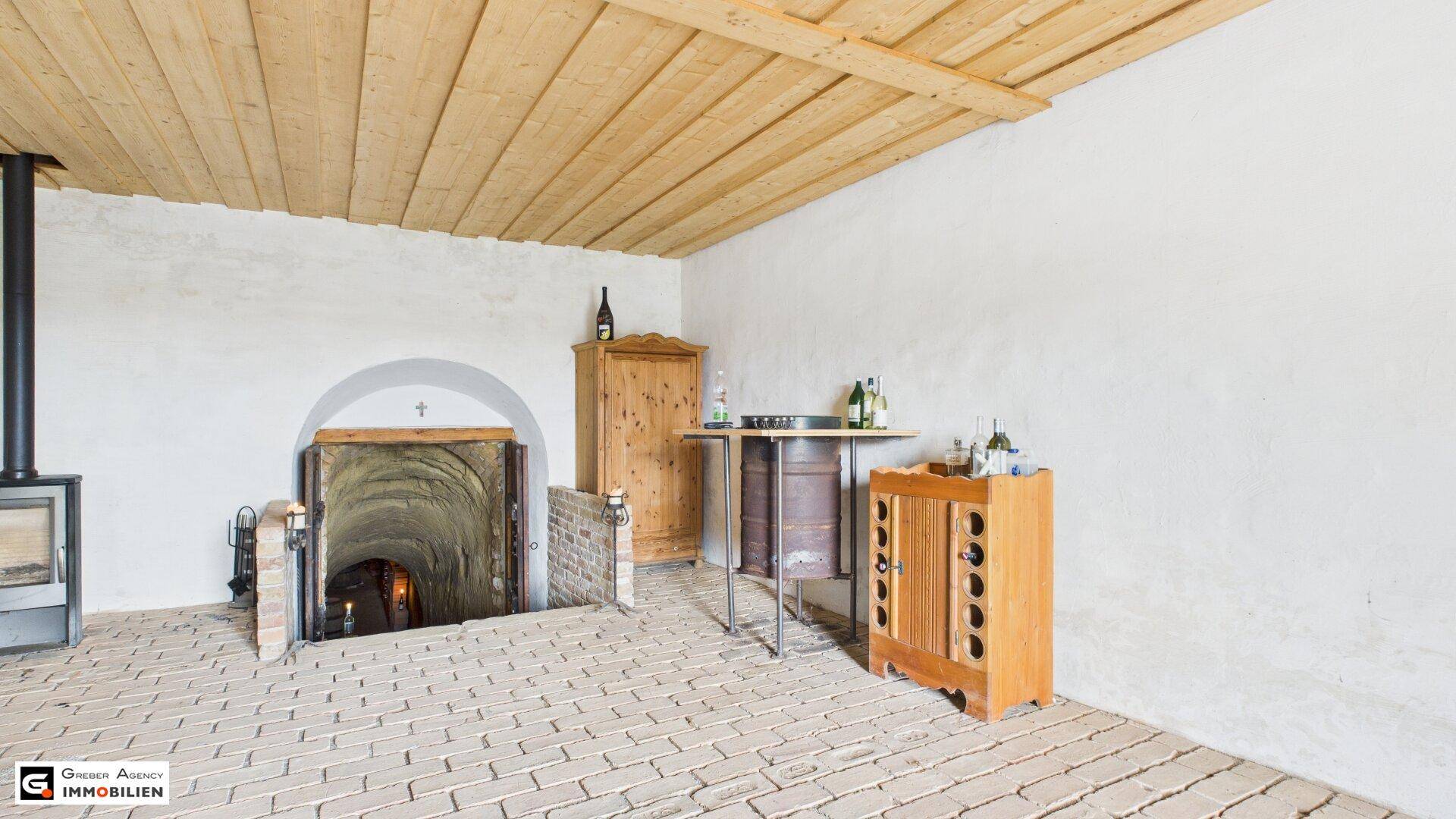 Rustikaler Eingangsbereich zum Weinkeller mit Holzbalkendecke, Ziegelboden, einem Kaminofen und Weinflaschen.