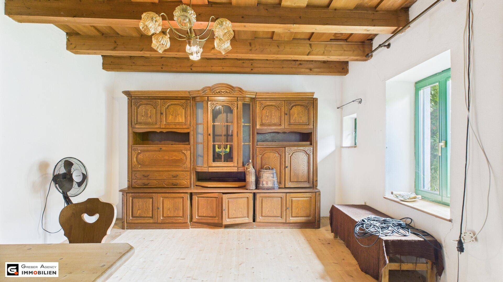 Geräumiger Wohnbereich mit großem Holzschrank, Holzbalkendecke und Blick ins Grüne durch das Fenster.