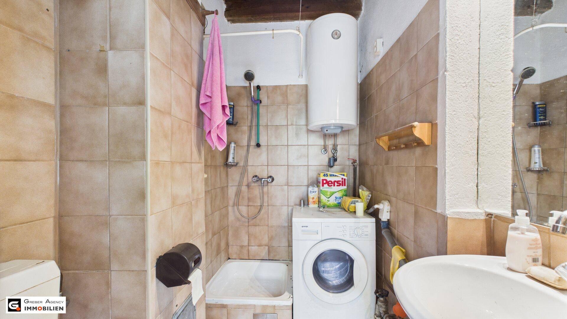Funktionales Badezimmer mit beigefarbenen Fliesen, Dusche, Waschmaschine und Warmwasserboiler.