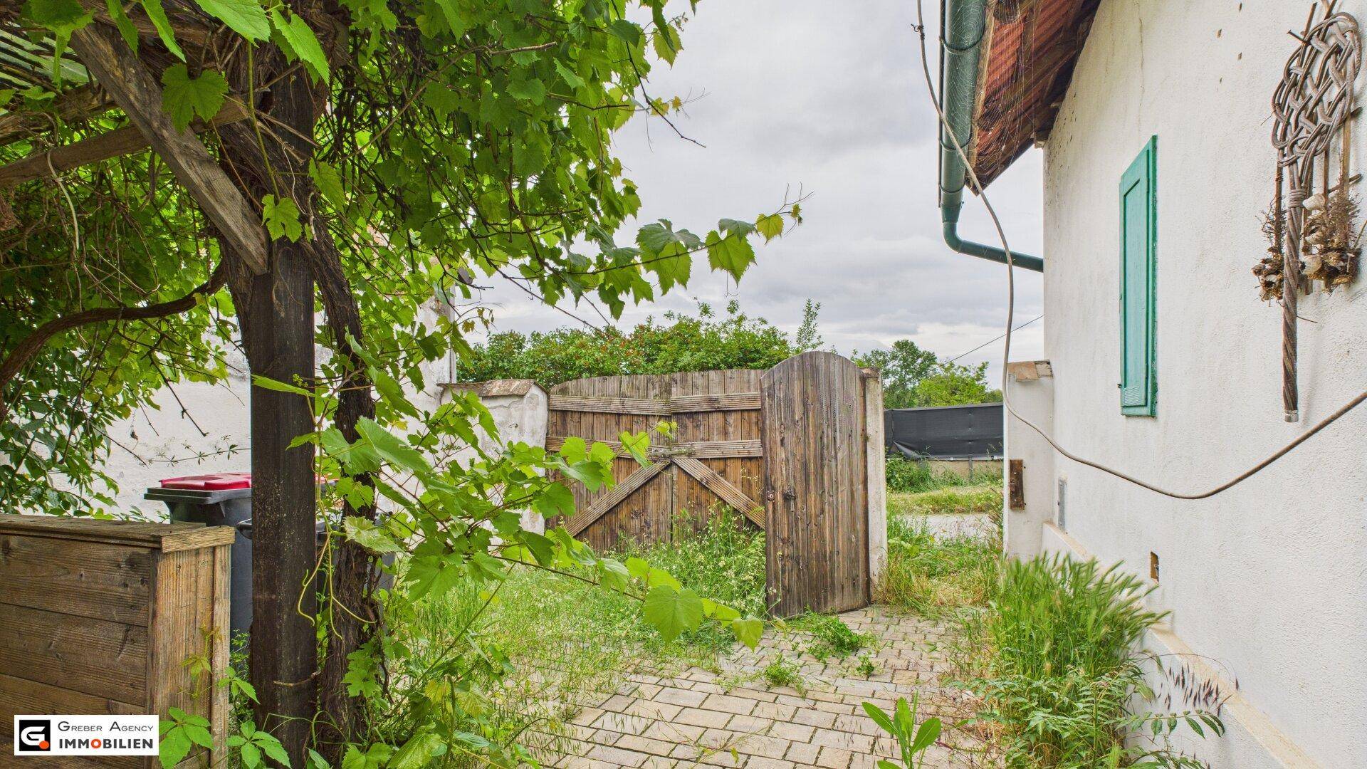 Gepflasterter Weg neben dem Haus, der zu einem hölzernen Gartentor und üppiger Vegetation führt.