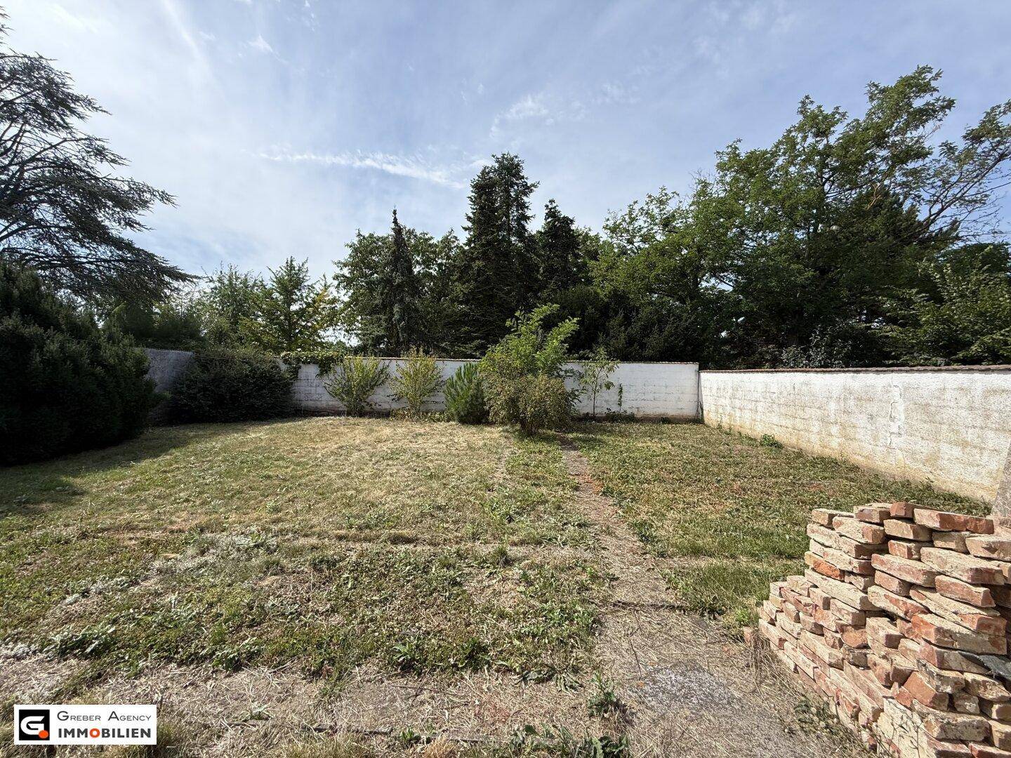 Weitläufiger Garten mit Rasenfläche, hohen Bäumen und einer Mauer im Hintergrund.