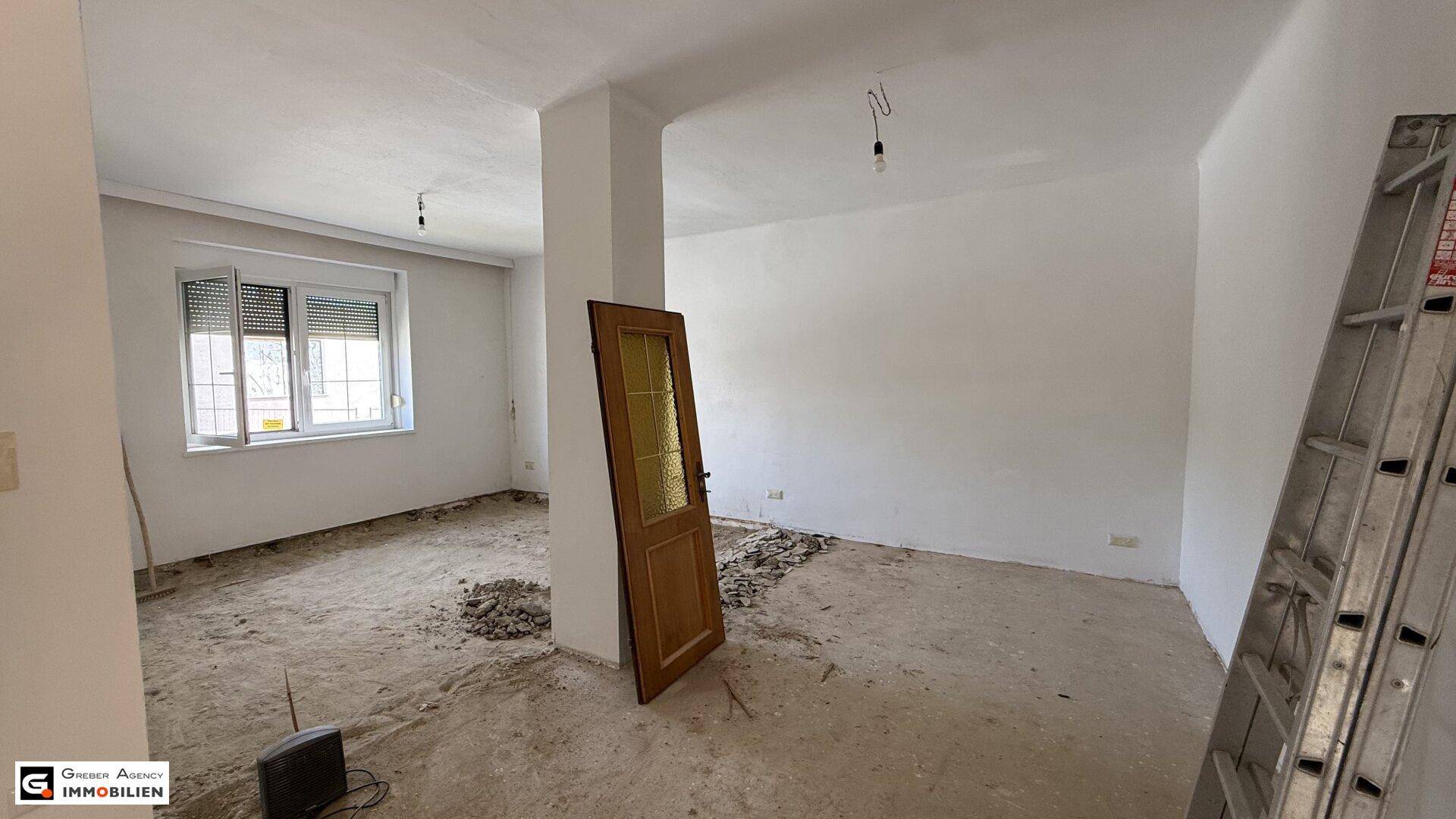 Innenraum im Rohzustand mit entfernten Bodenbelägen und einem Fenster mit Rollladen.