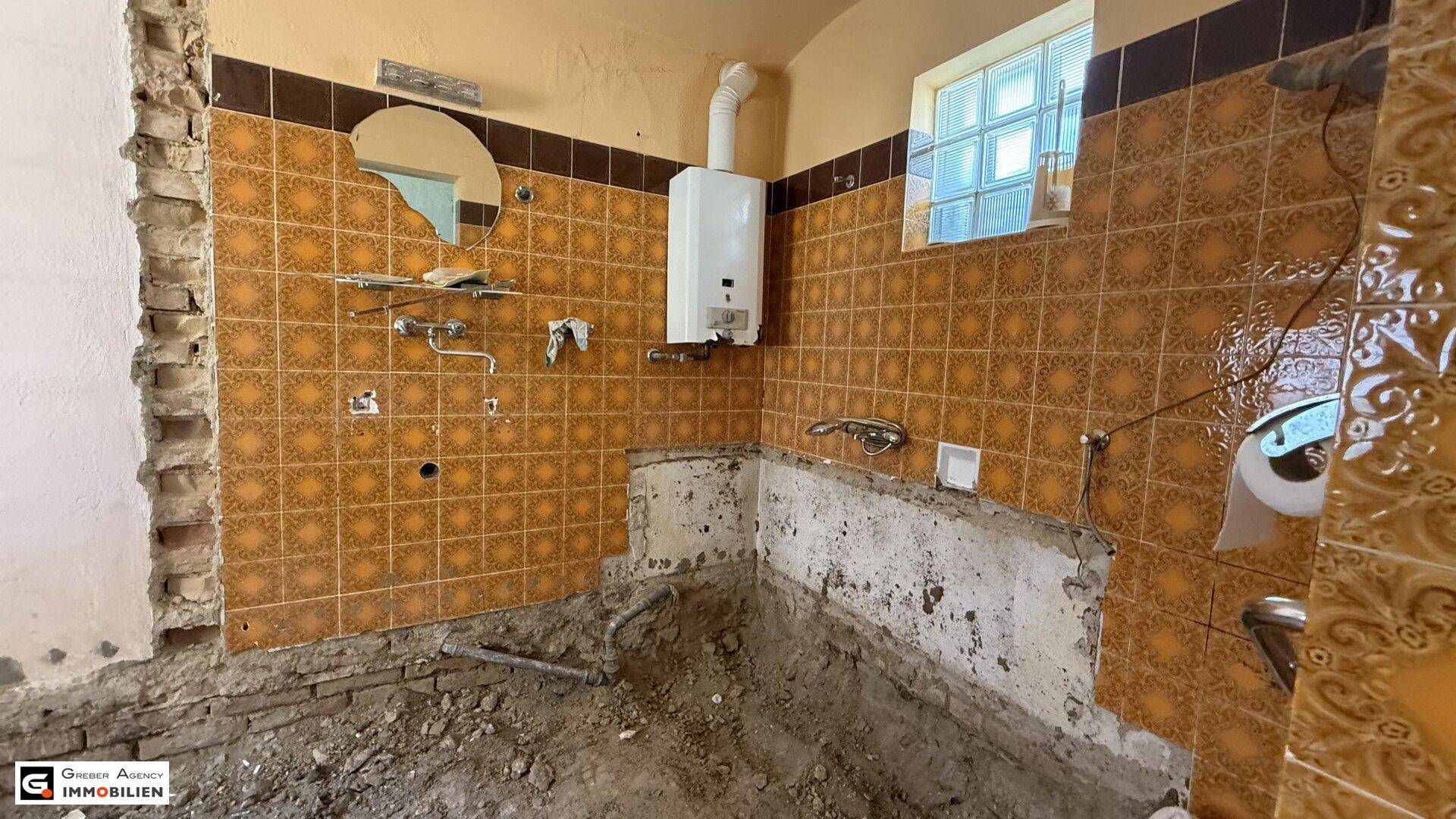 Badezimmer im Rohbau mit alten Fliesen, Gastherme und einem kleinen Fenster.