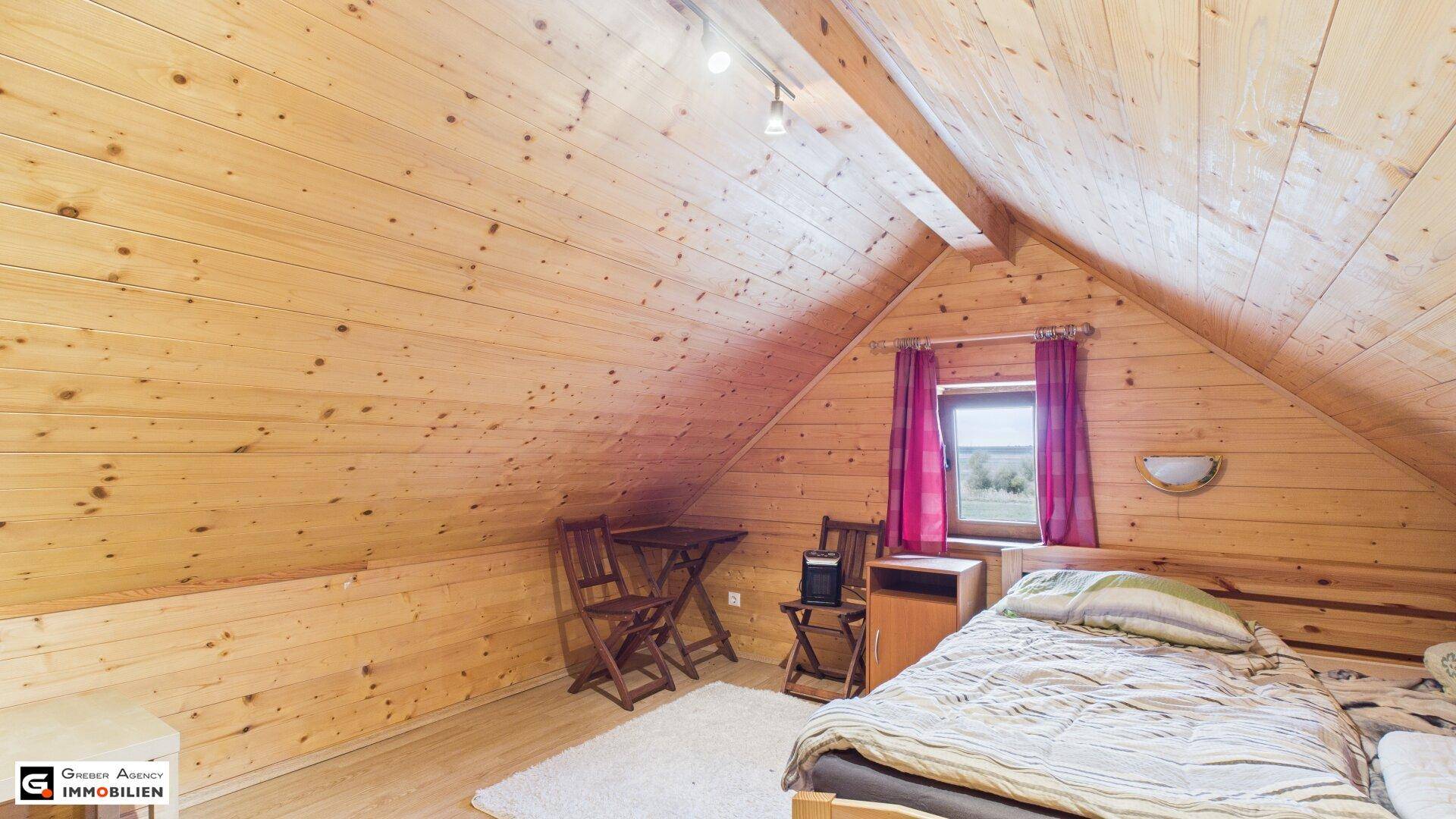 Holzvertäfeltes Schlafzimmer im Dachgeschoss mit schrägen Wänden, Fenster und einfacher Einrichtung.